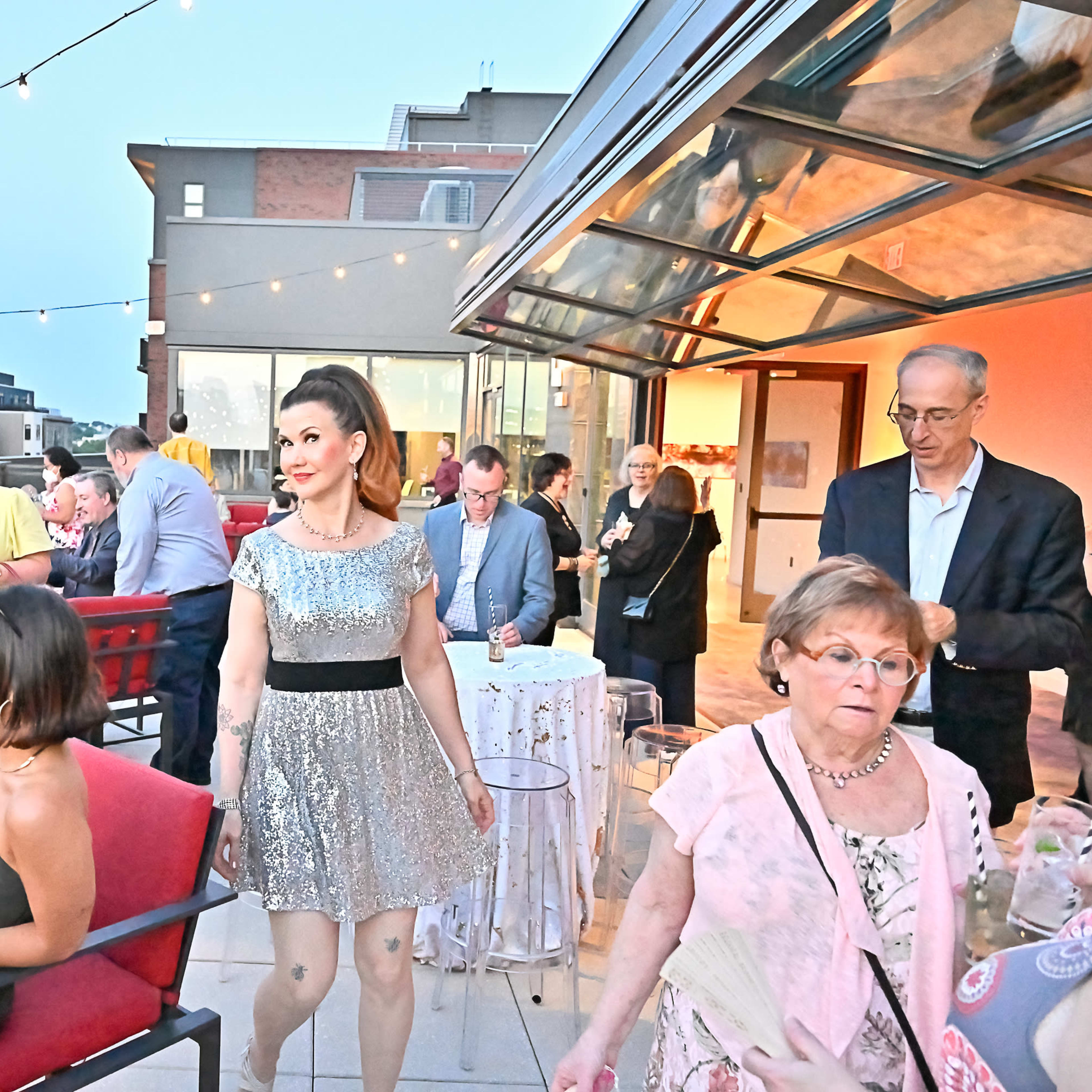 A group of elegantly dressed people socializes on a rooftop terrace, surrounded by string lights and modern urban buildings in the background.