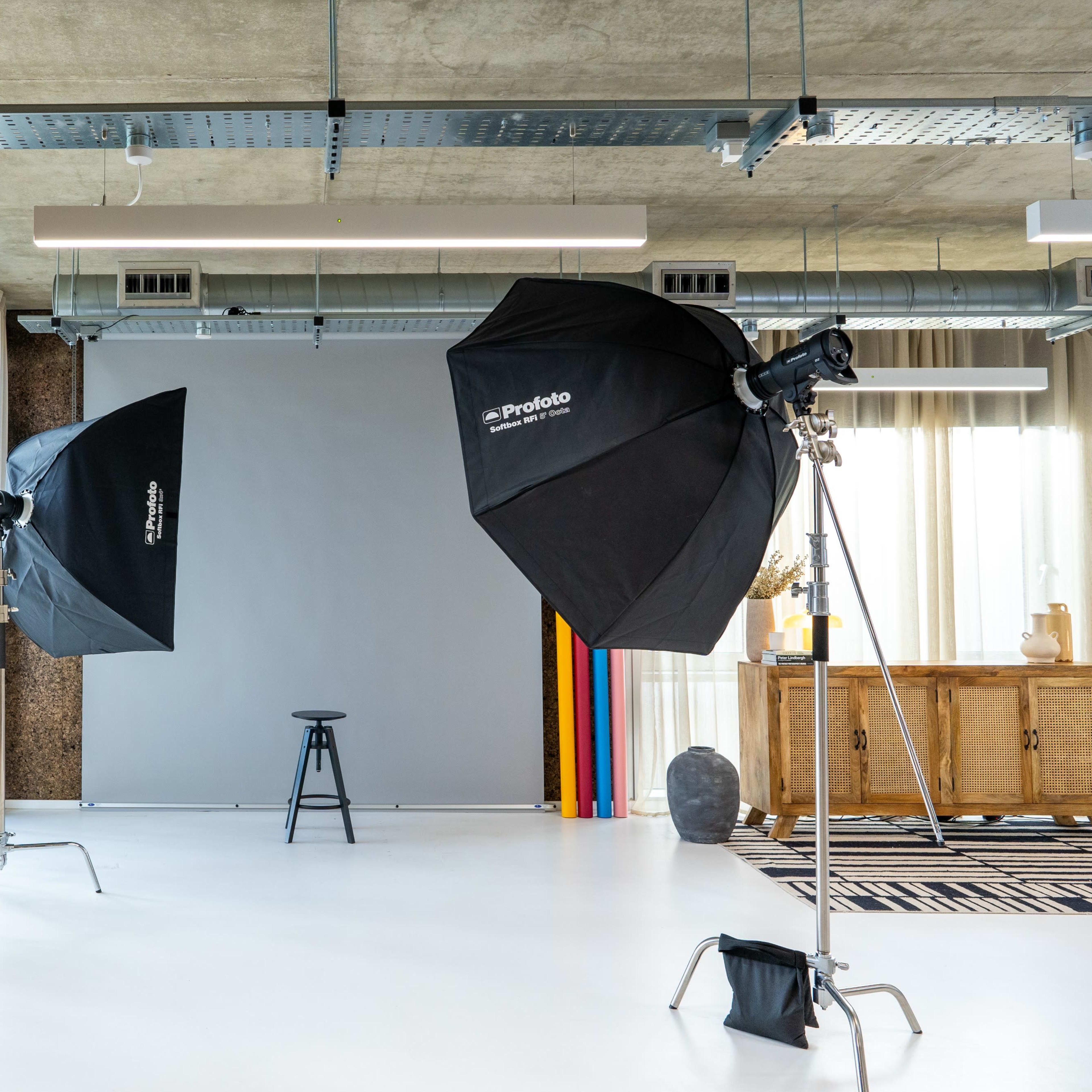A photography studio is set up with two softbox lights, a gray backdrop, and a stool in the center, surrounded by large windows with sheer curtains.