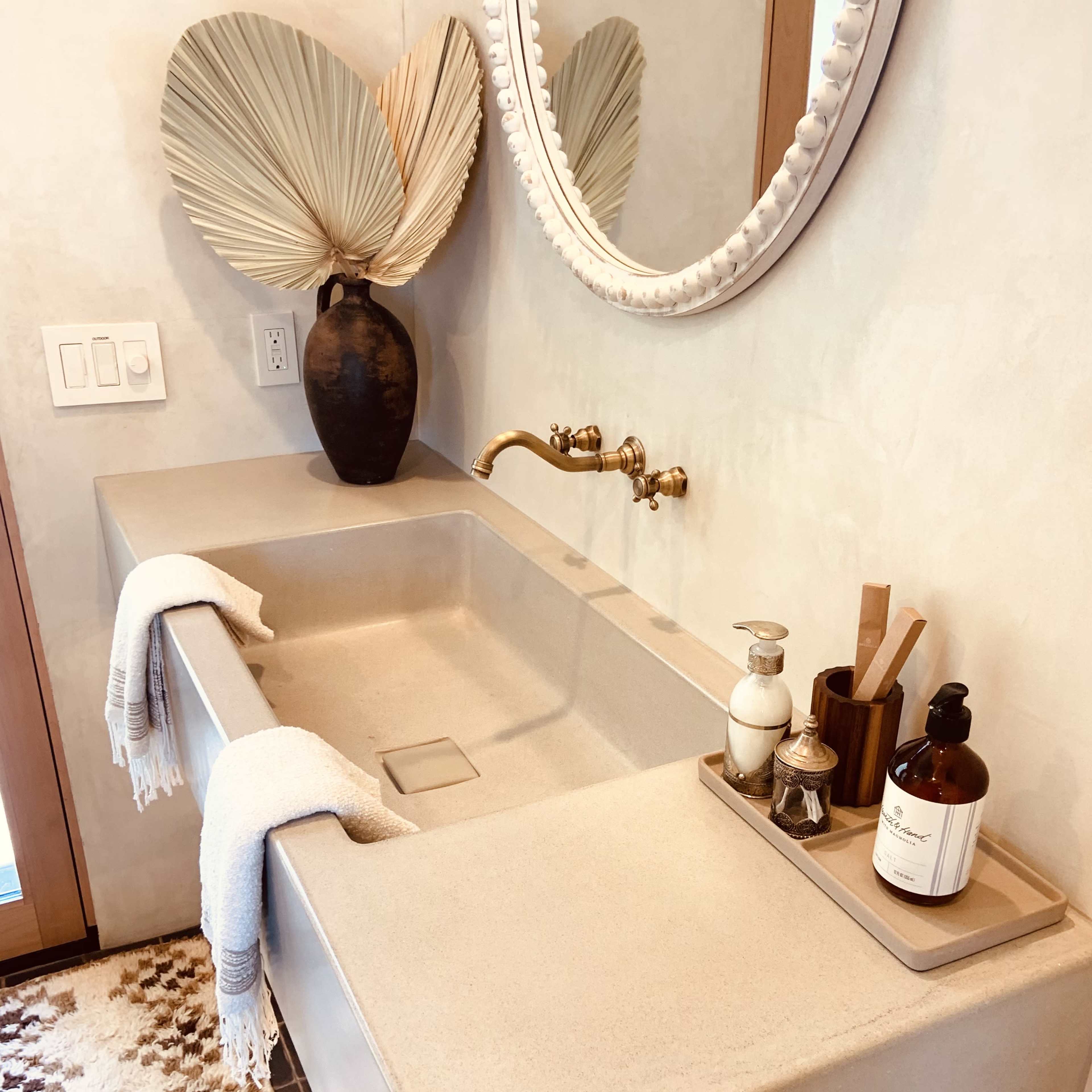 A modern bathroom features a large, rectangular sink with a wall-mounted faucet, a round mirror, decorative palms, and various toiletries displayed on a tray.