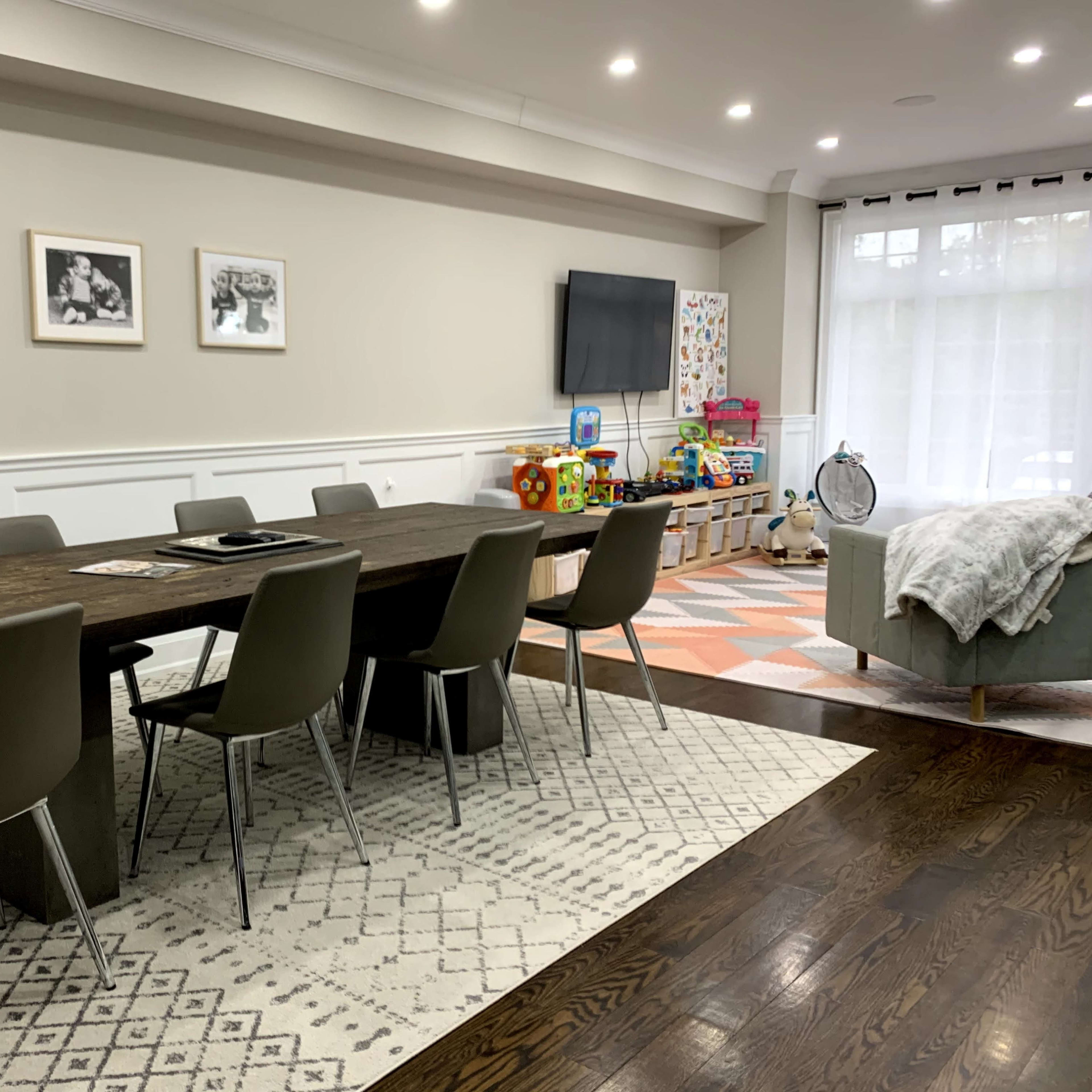 A dining area features a long wooden table surrounded by gray chairs, with a television mounted on the wall and colorful toys positioned nearby.