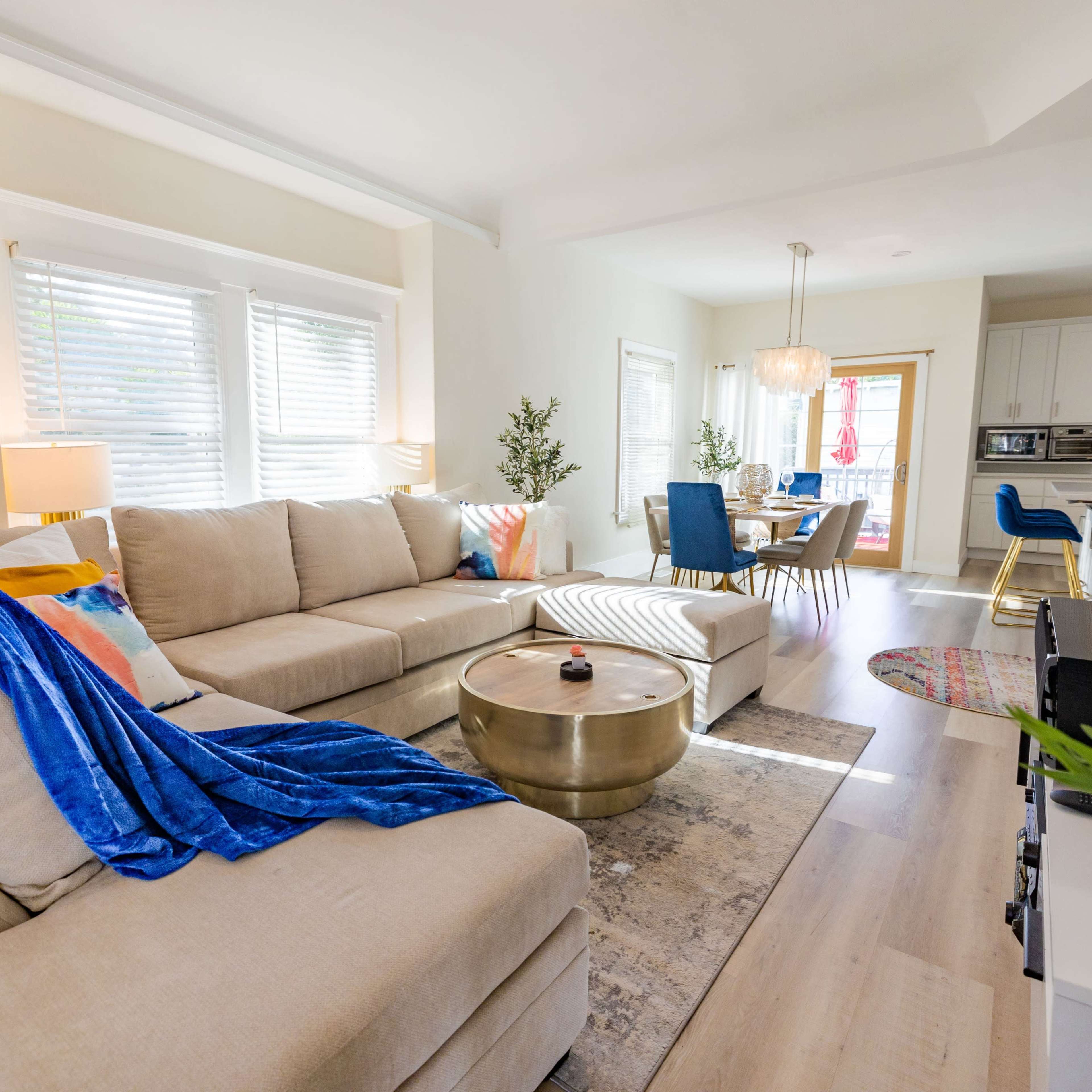 A well-furnished living room with a beige sectional sofa, a round metal coffee table, and a dining area visible in the background.