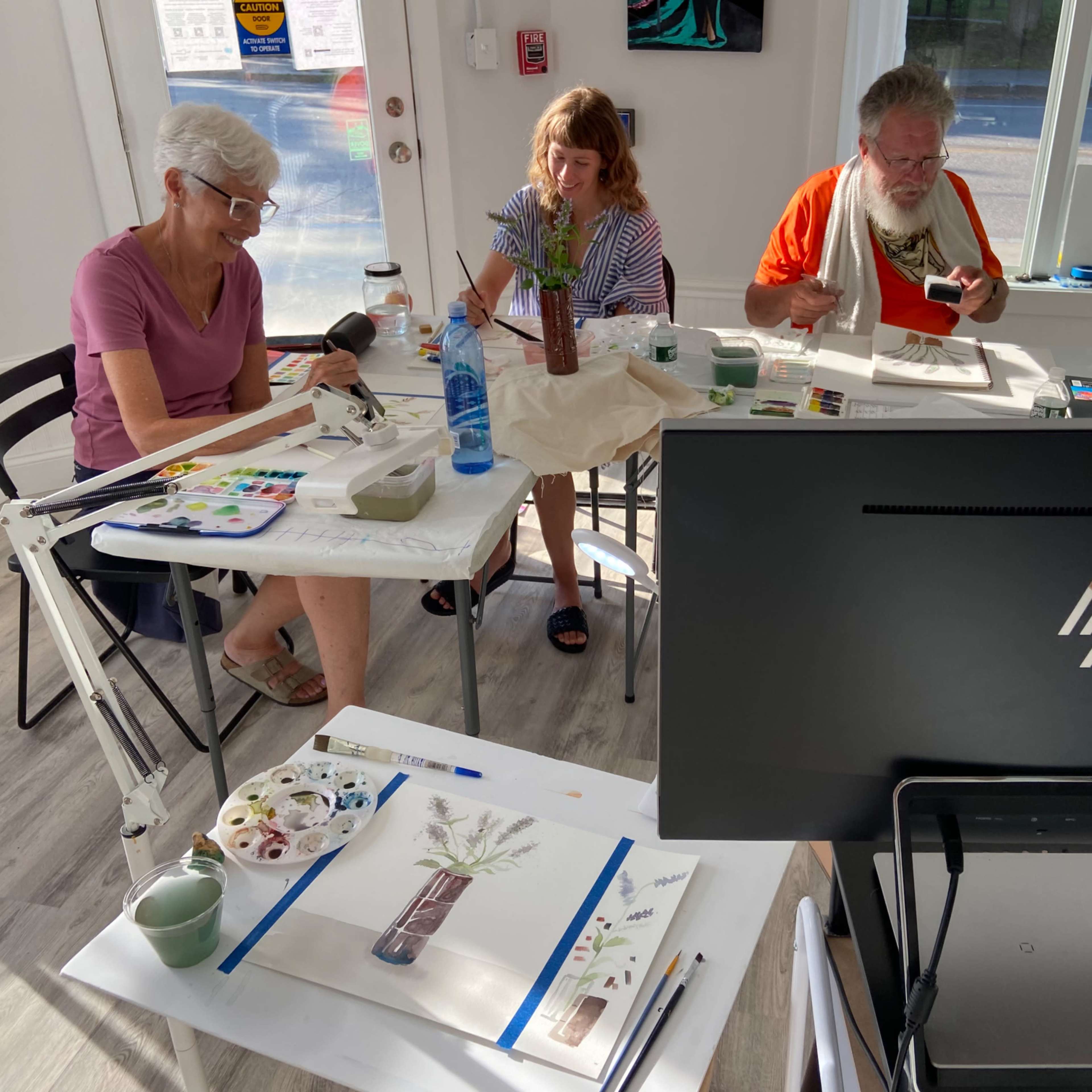 Three people are seated at a table engaged in a painting class, surrounded by art supplies and natural light coming through a large window.