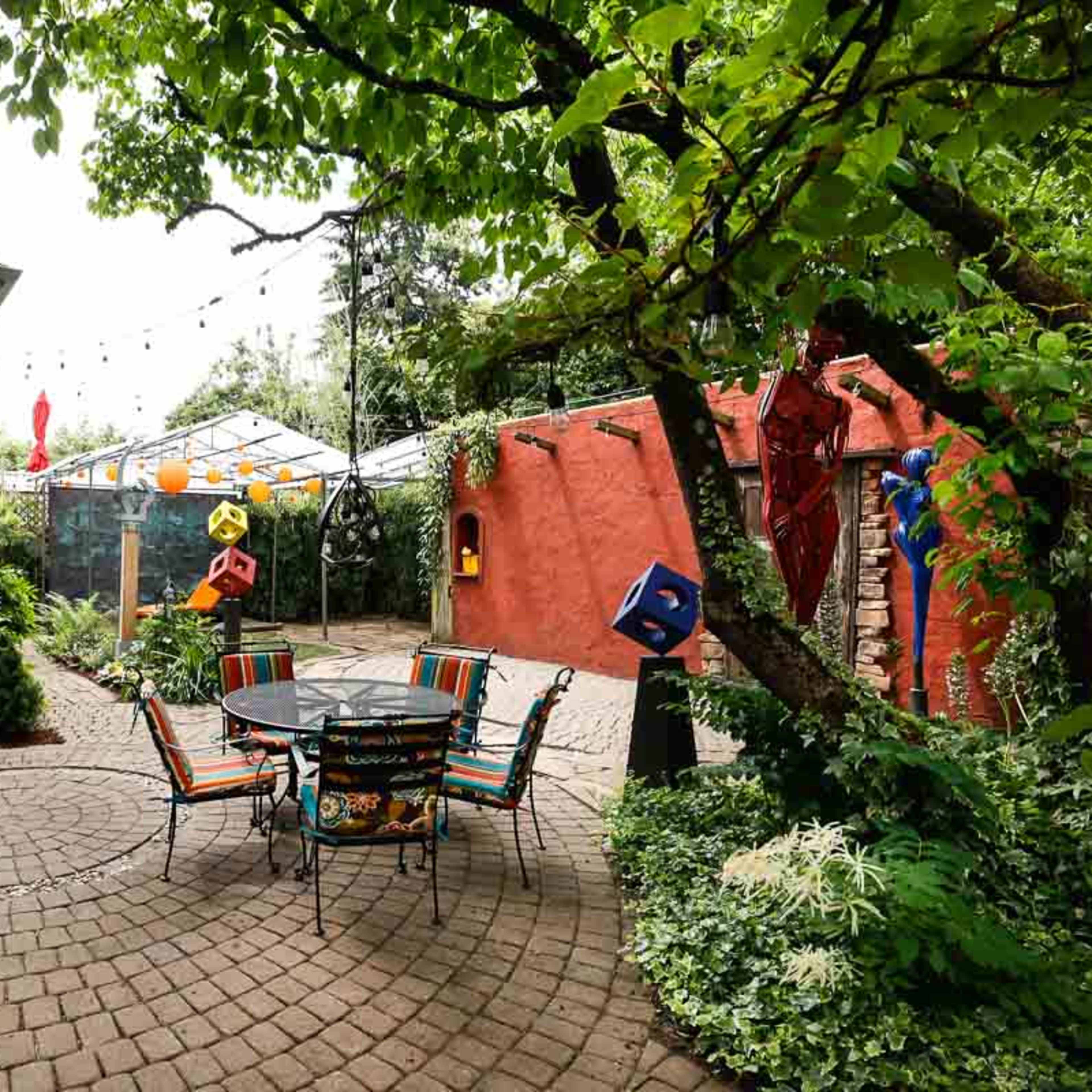 A circular stone patio features a black metal table and chairs surrounded by lush greenery and colorful decorations.
