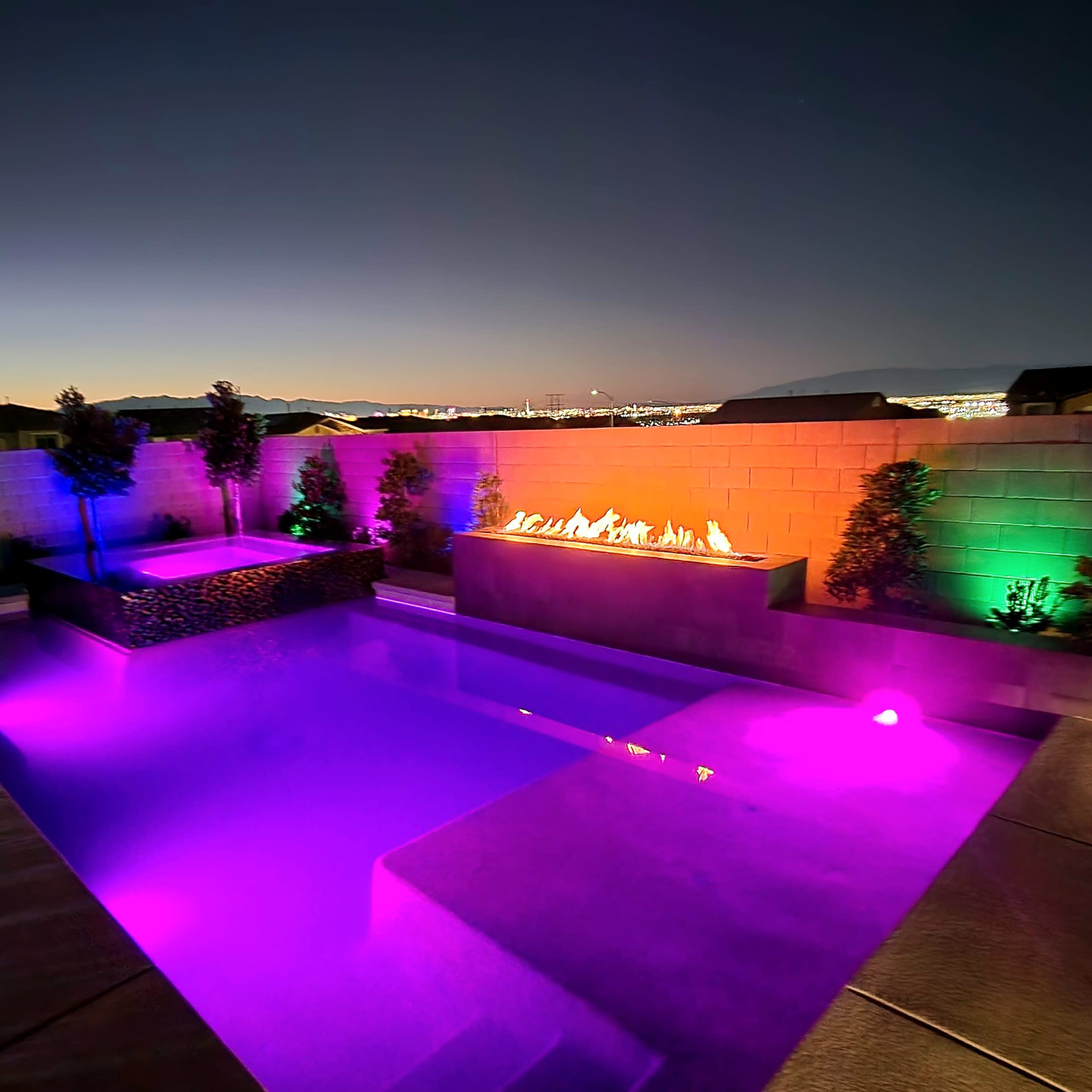 A swimming pool at night, illuminated by colorful lights and featuring a fire feature along a stone wall.