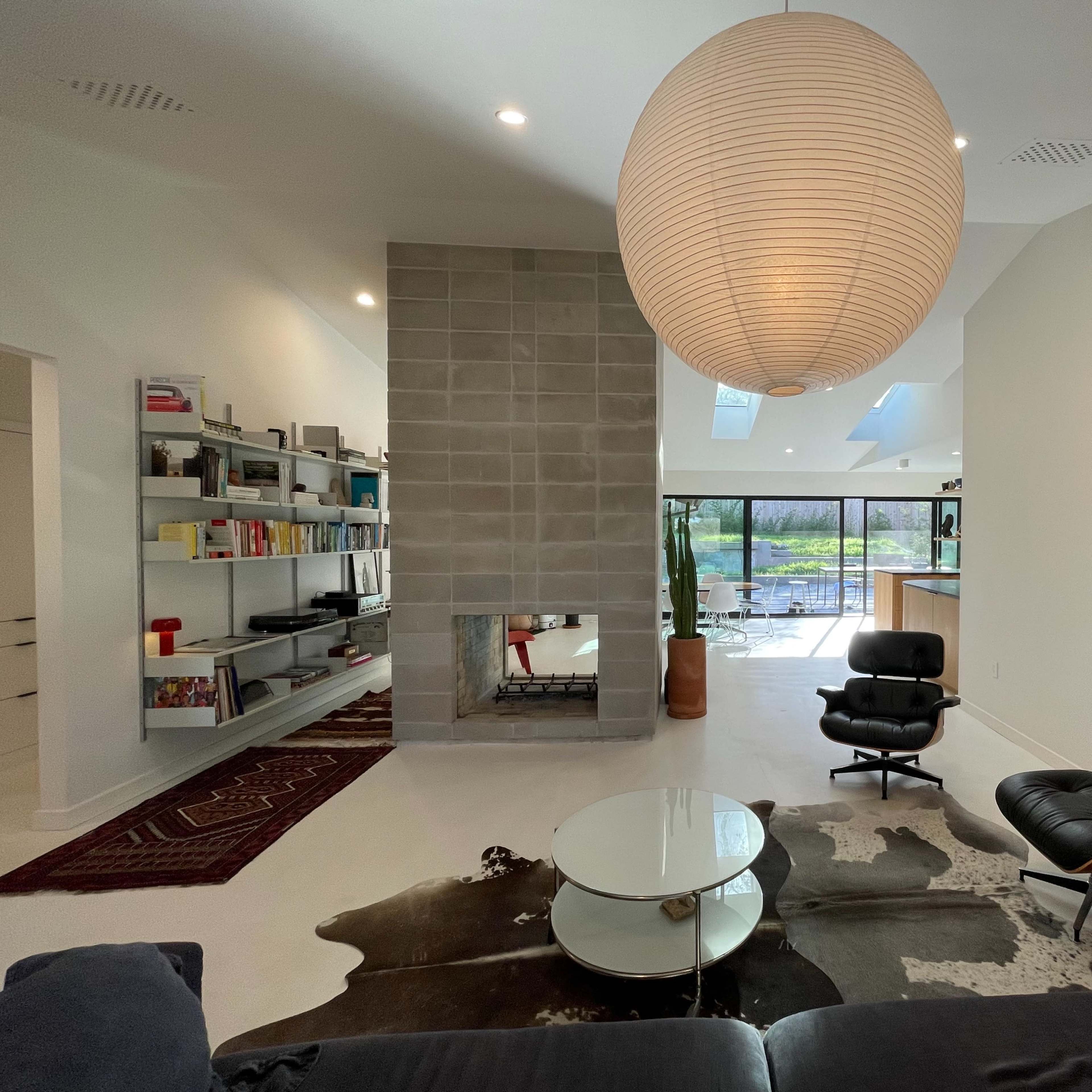 A modern living room with a large round pendant light, a black lounge chair, a cowhide rug, a stone fireplace, and a view of a courtyard through large glass doors.