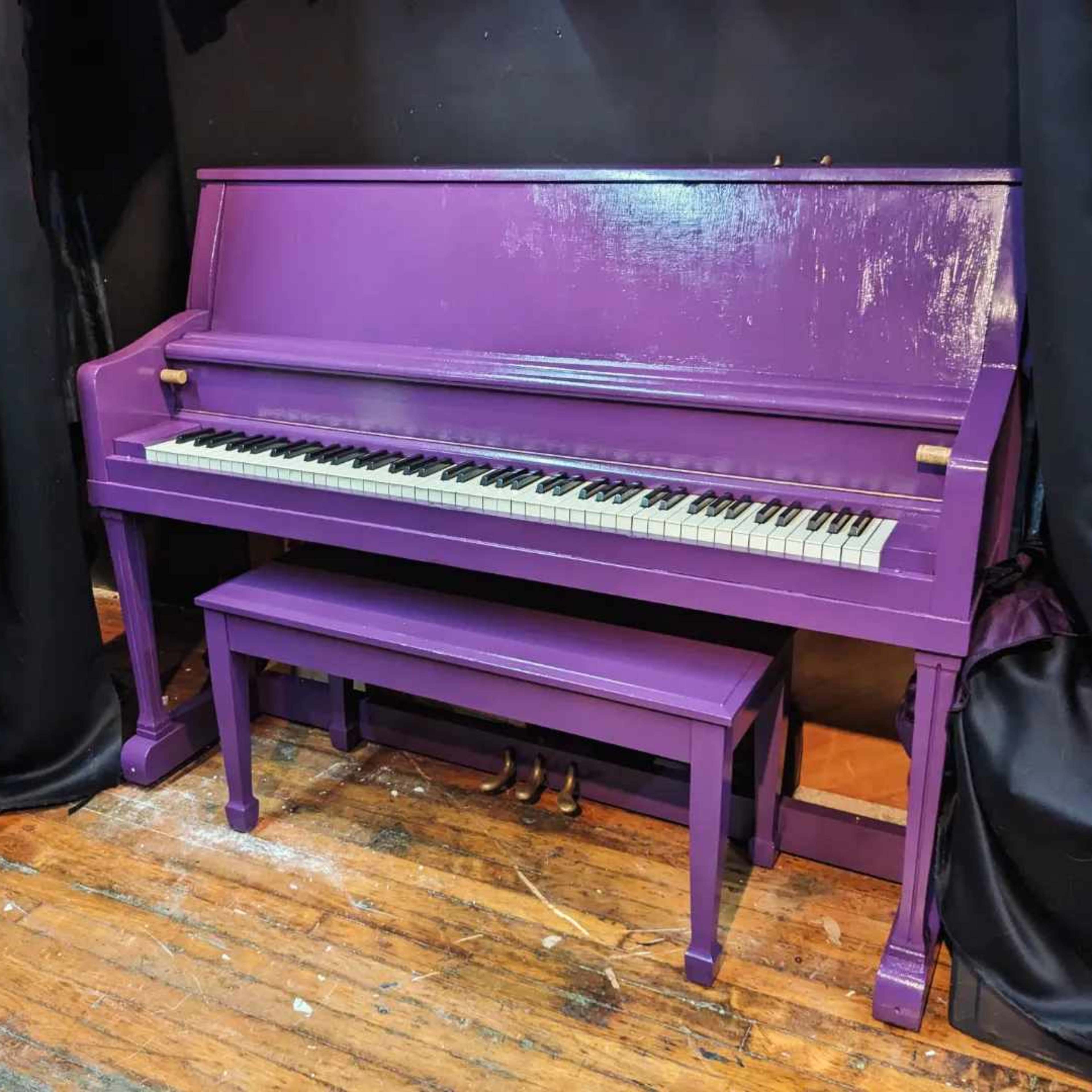 A purple piano and matching bench are positioned against a black backdrop in a dimly lit room.