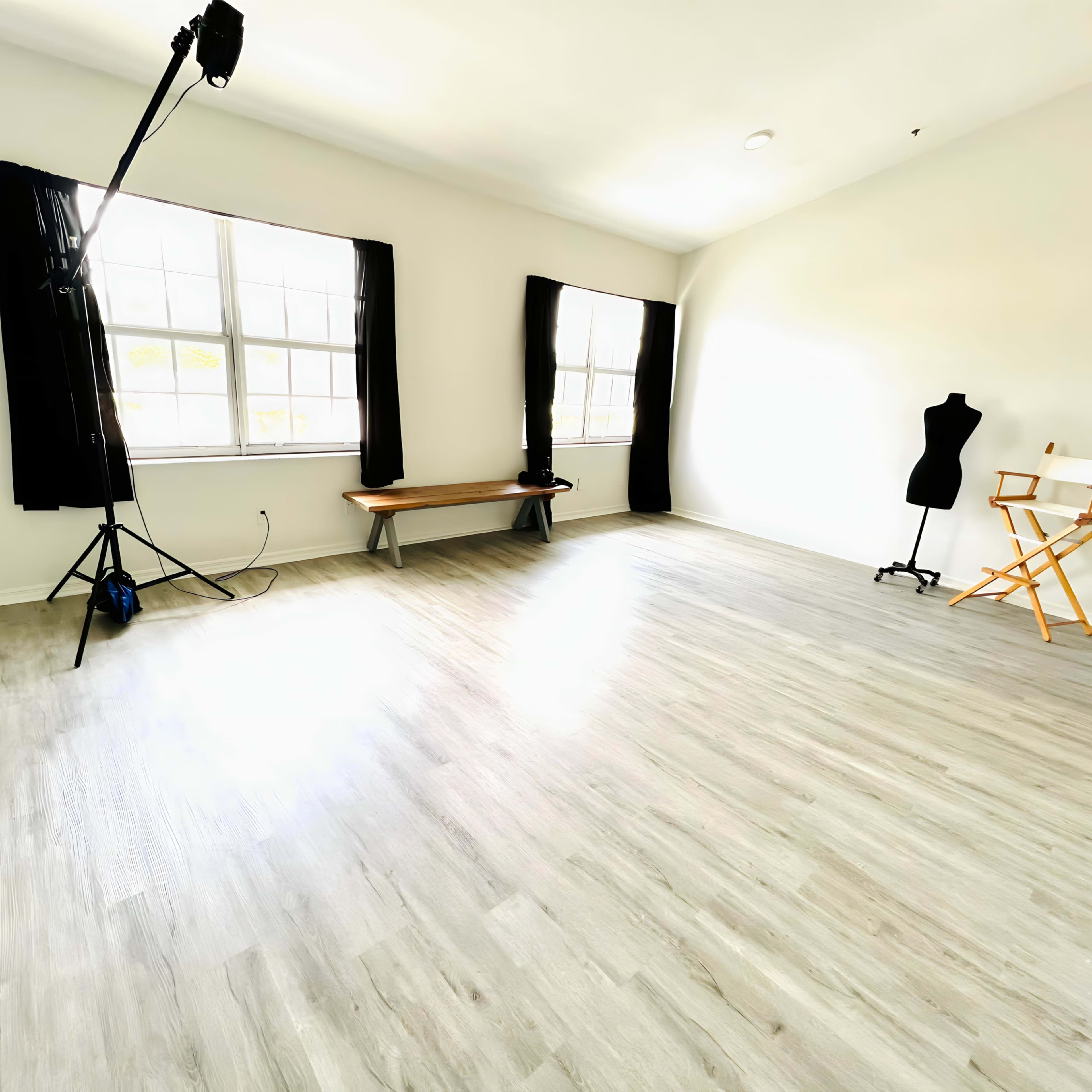 A well-lit room with light-colored flooring, two large windows with black curtains, a wooden bench, a chair, and a dress form.