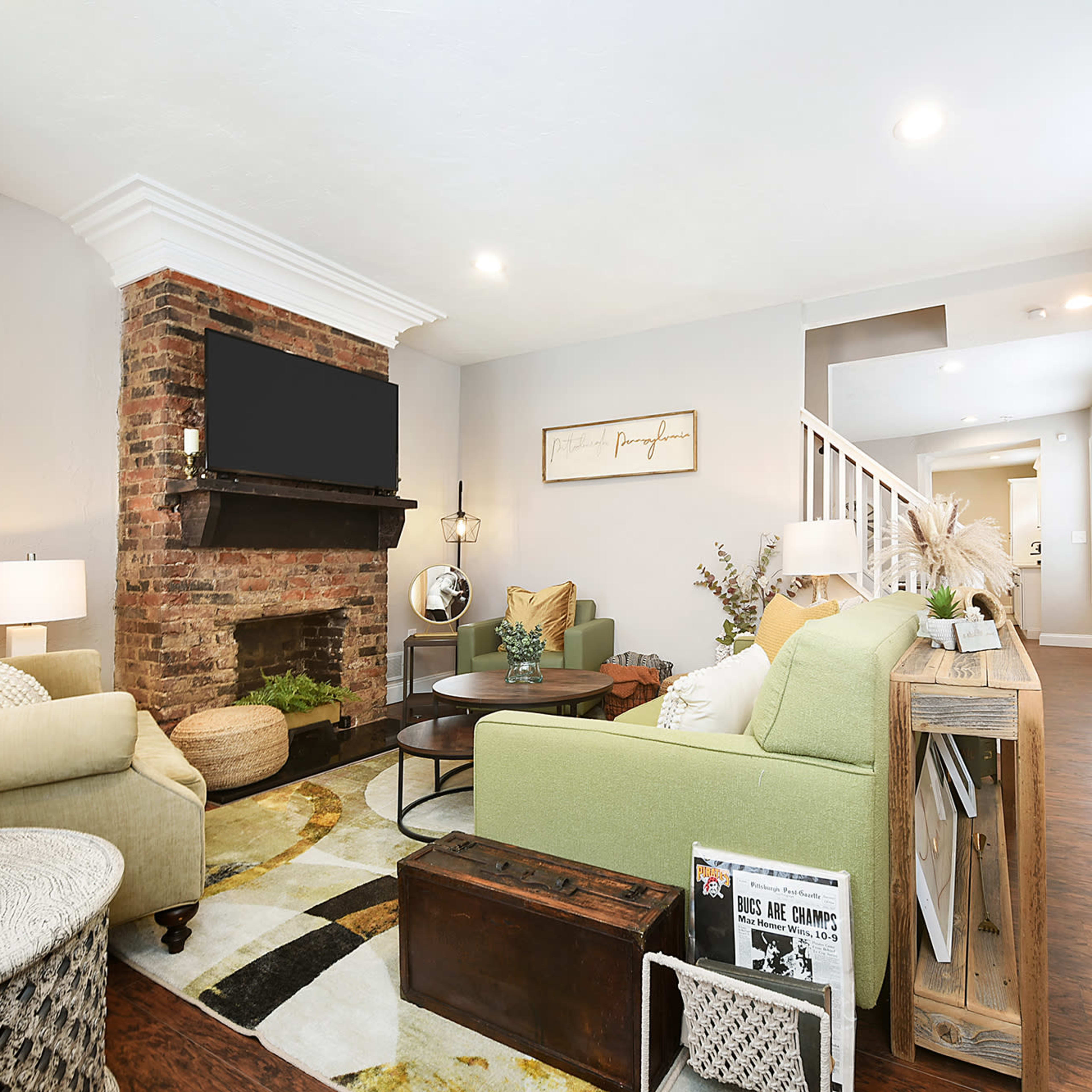 The living room features a brick fireplace with a mounted TV, a green sofa, and a round coffee table surrounded by decorative elements and natural light from the windows.