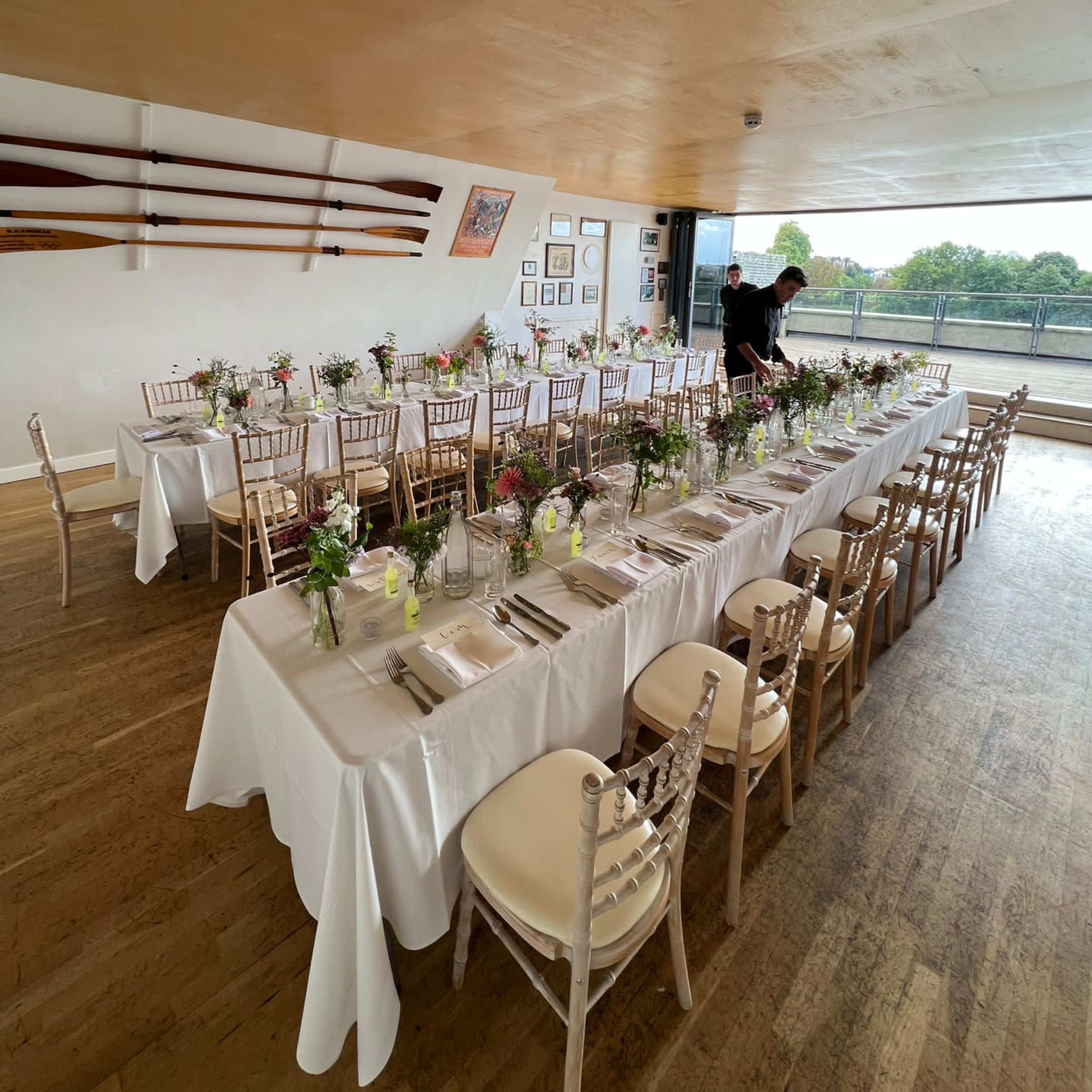 A long table set with white tablecloths and floral centerpieces is arranged in a dining room with large windows overlooking a view.