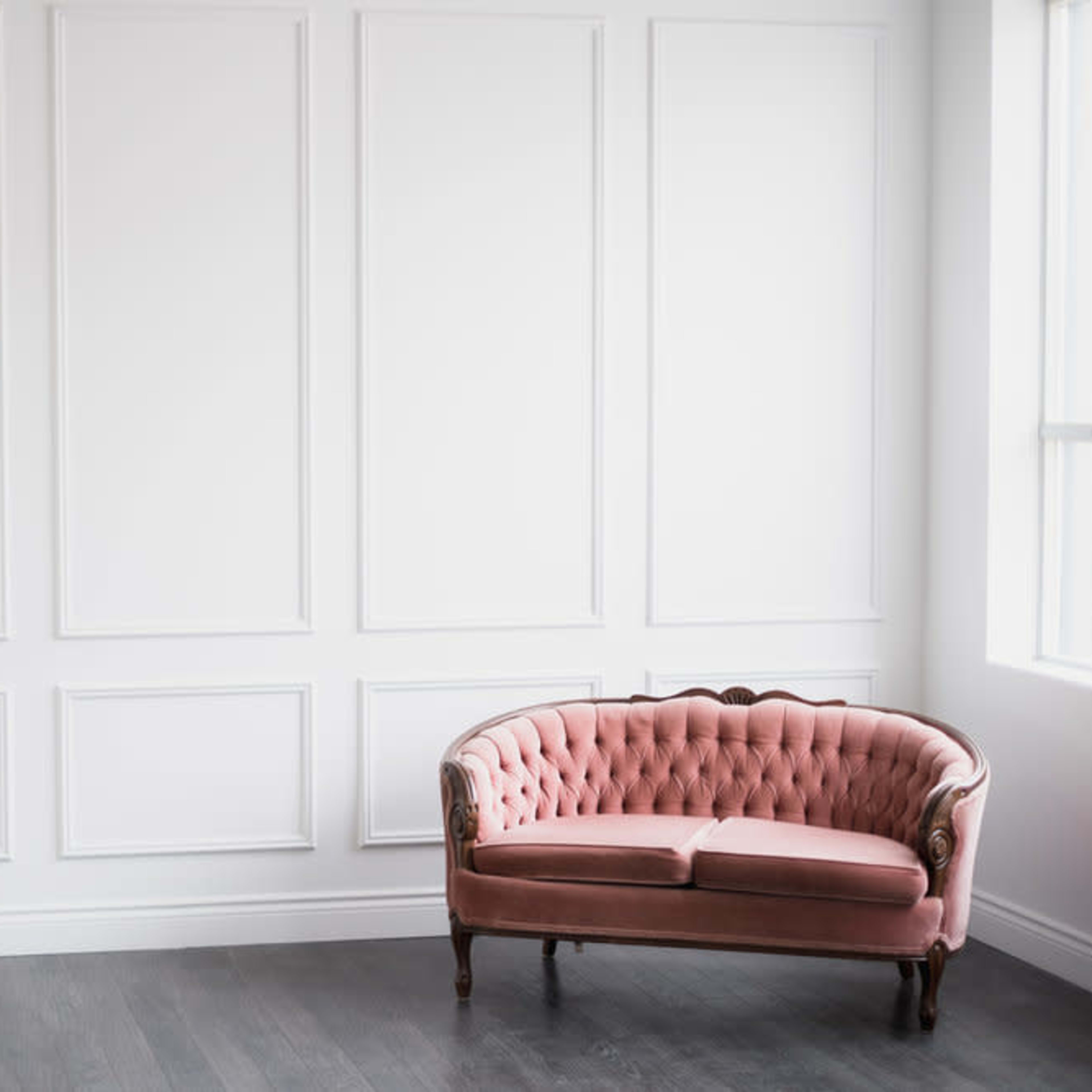 A tufted pink sofa is positioned against a white paneled wall in a brightly lit room.