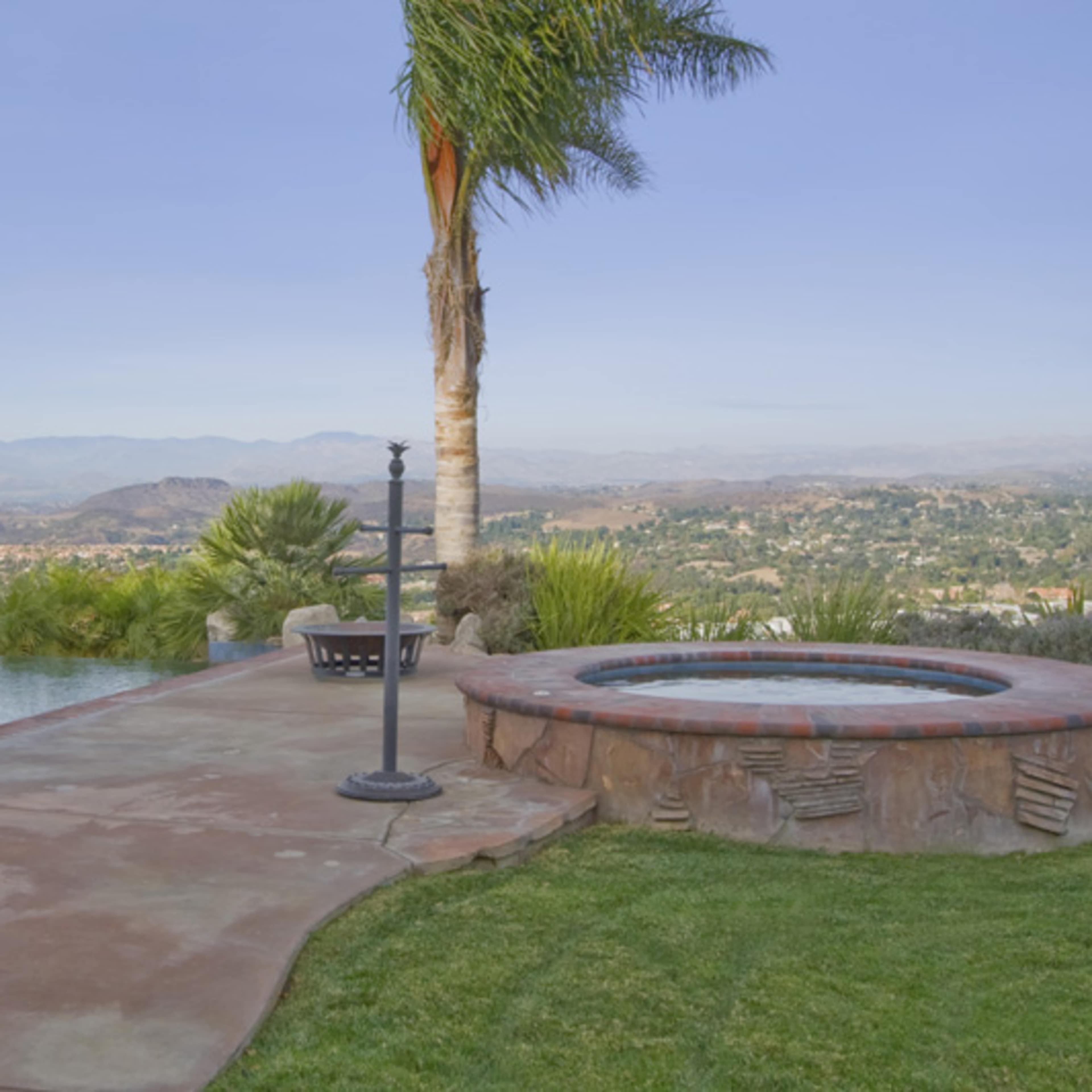 A backyard scene features a swimming pool adjacent to a hot tub, with a view of rolling hills and a clear sky in the background.