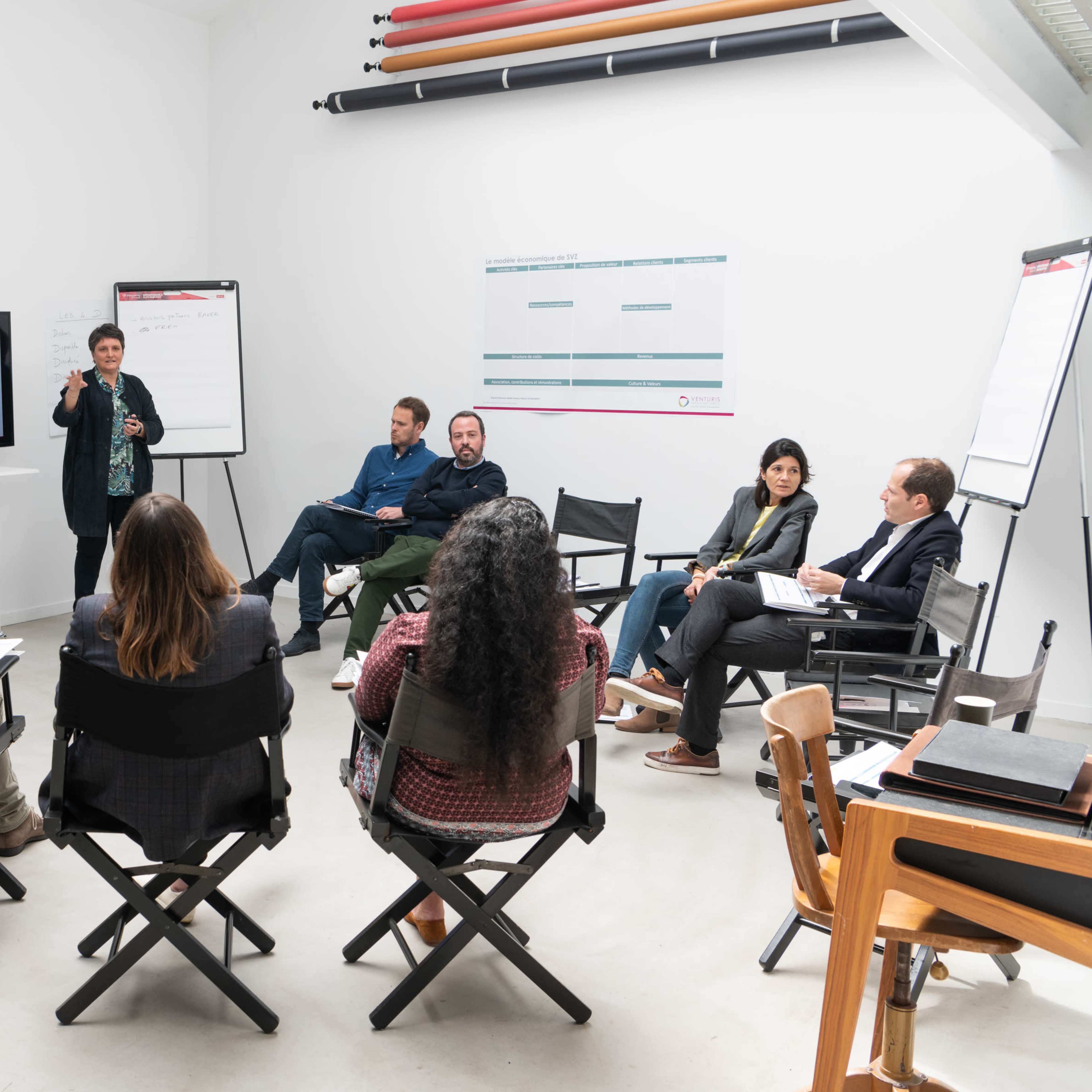 A group of eight people is seated in a circular arrangement in a well-lit room, with a presenter standing and pointing at a screen while others engage attentively.