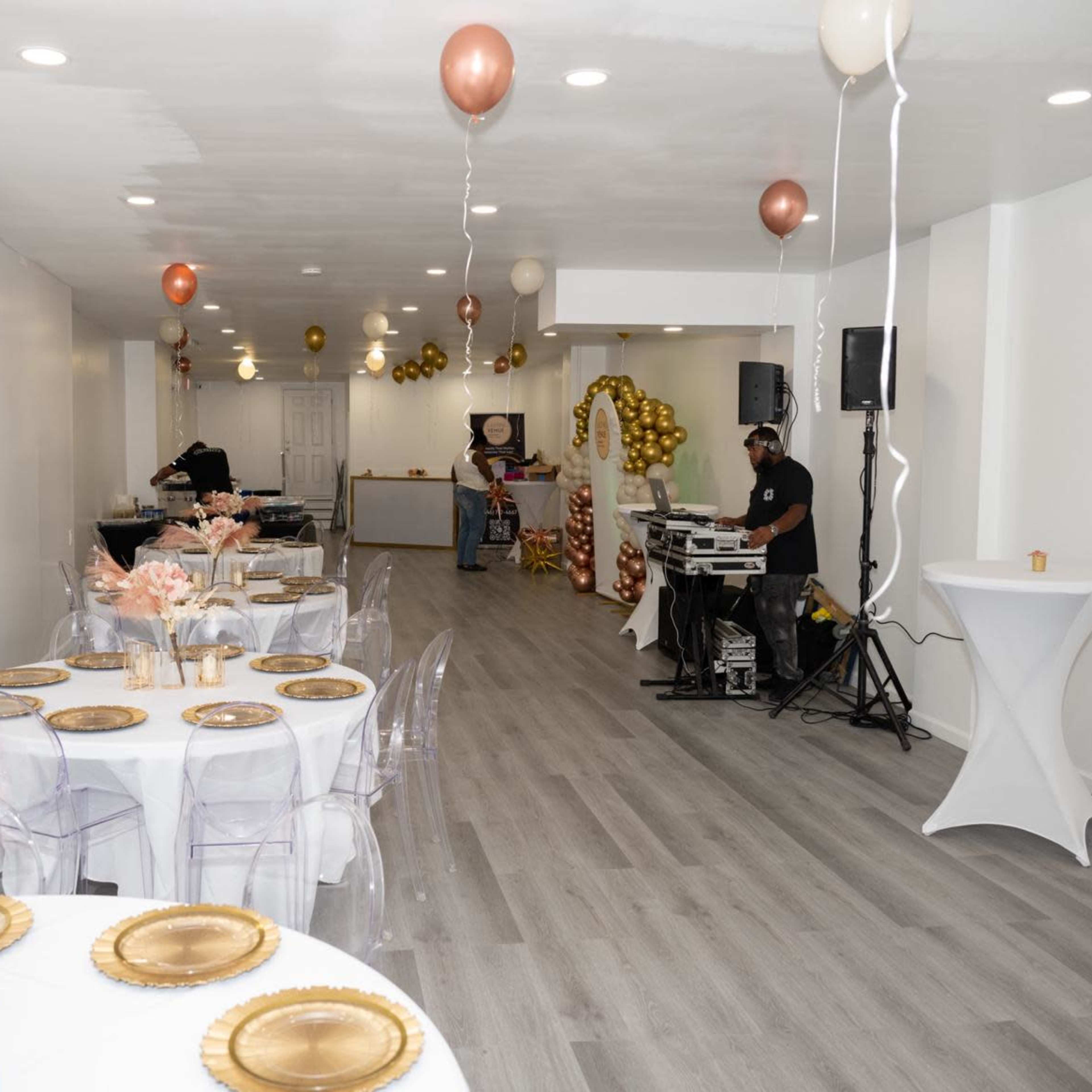 A banquet hall decorated with round tables, gold and rose gold balloons, and a DJ setup in the corner.