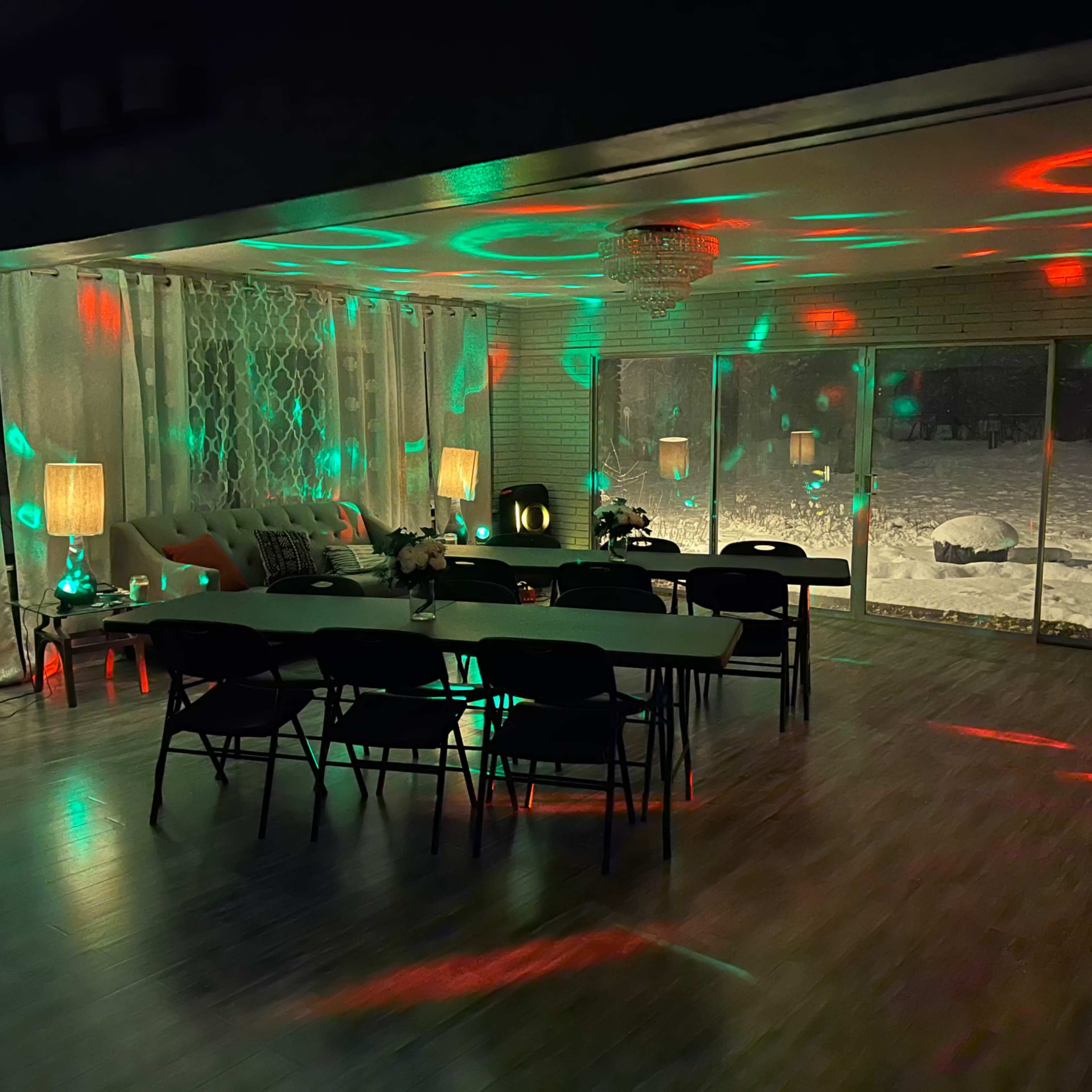 A brightly lit room features a long table surrounded by chairs, with colorful lights casting patterns on the walls and a snowy landscape visible through large windows.