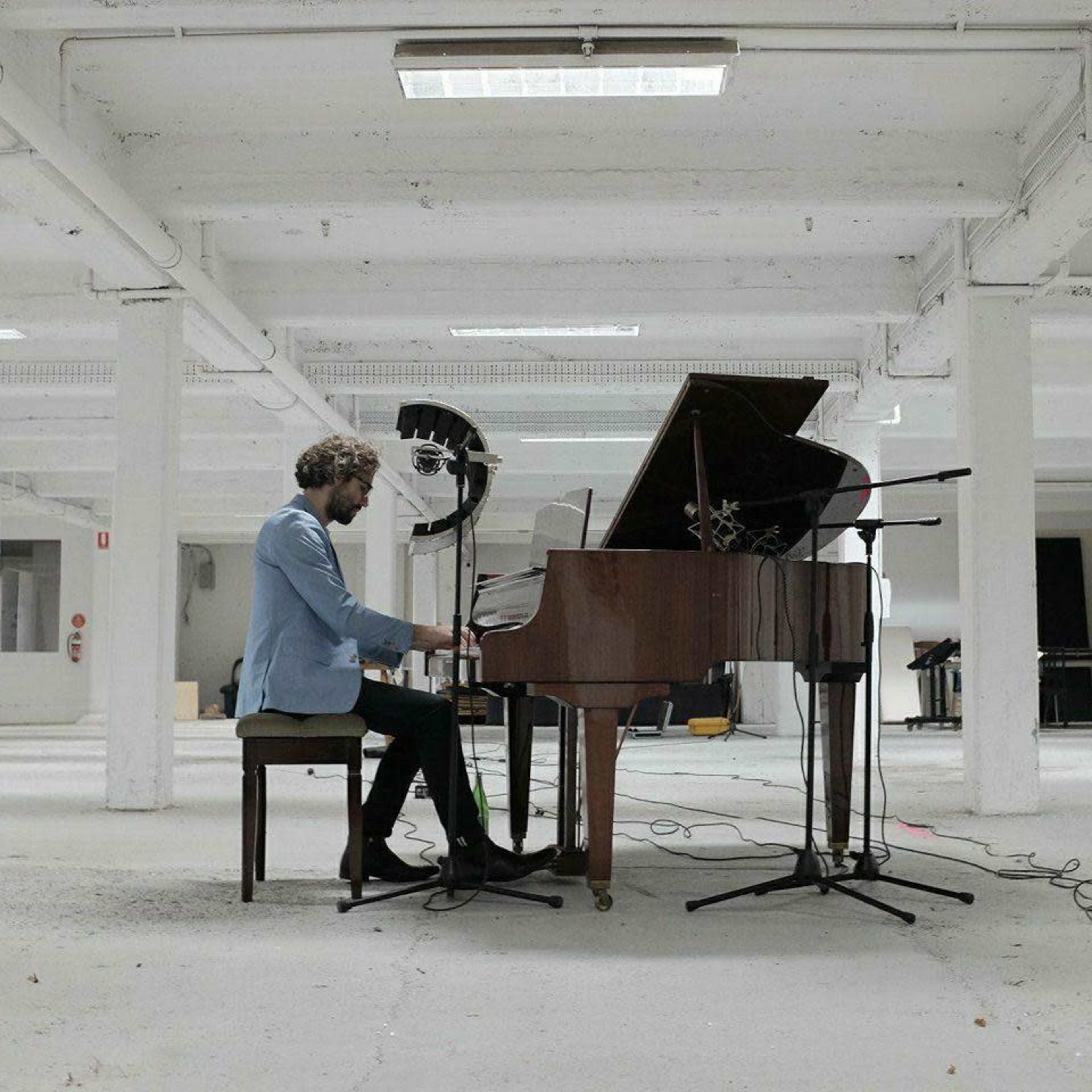 A musician plays a grand piano in a spacious, empty room with white walls and visible microphones set up around him.
