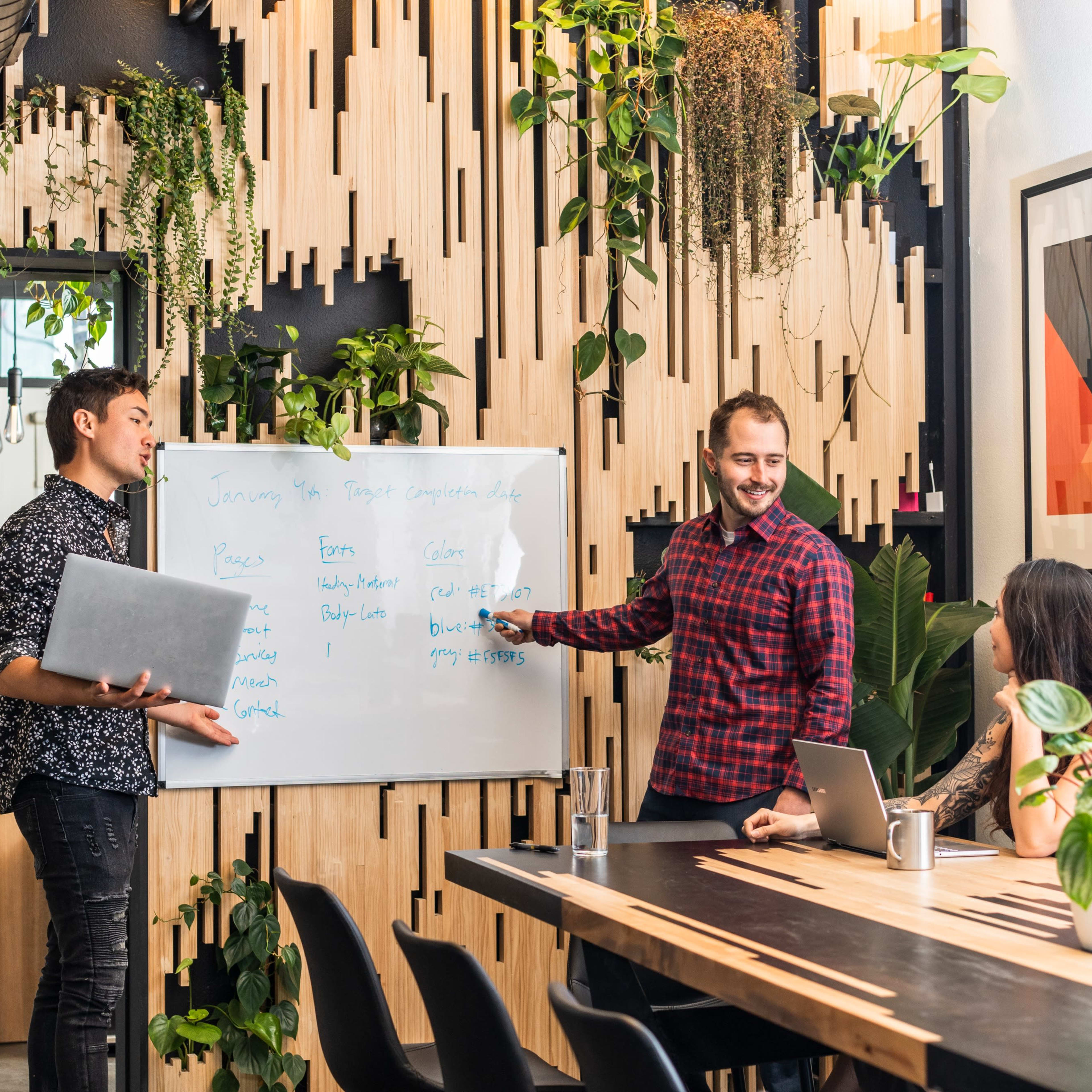 Three people engage in a meeting around a whiteboard in a stylish office space filled with plants and wooden accents.