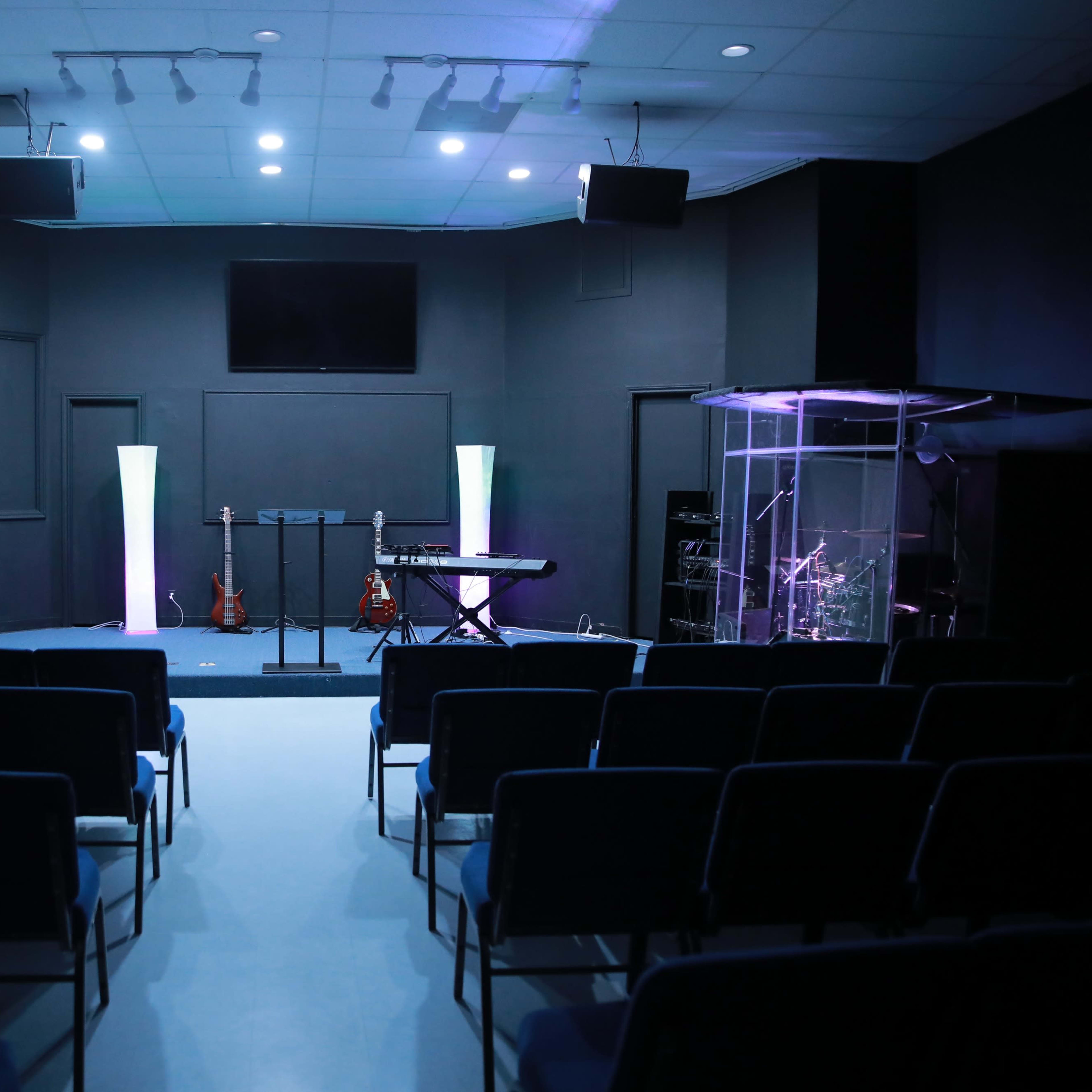 A small auditorium with rows of chairs facing a stage equipped with musical instruments and a transparent sound booth.