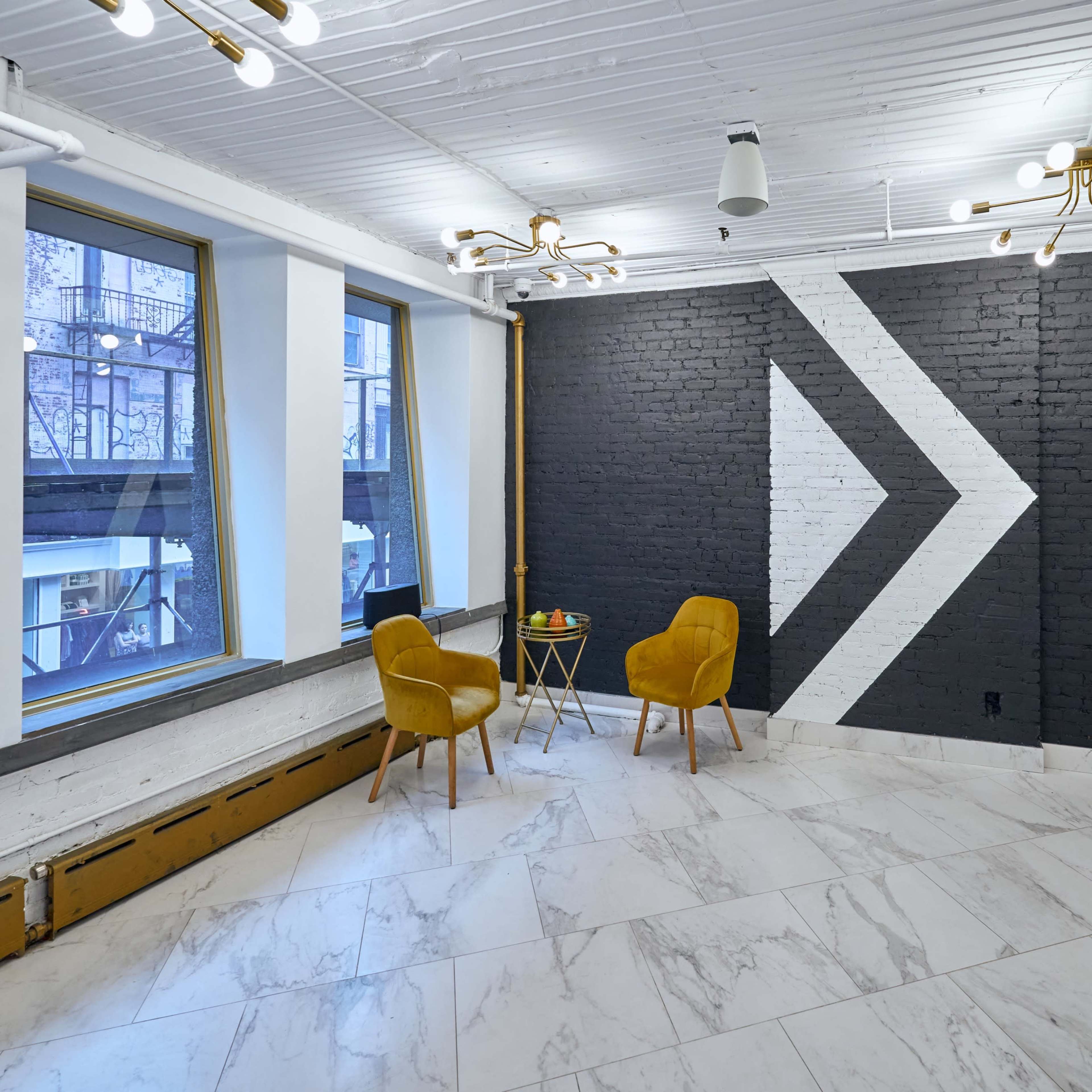 A modern interior space featuring a black and white accent wall with an arrow design, two yellow chairs, a small table with fruit, and large windows.