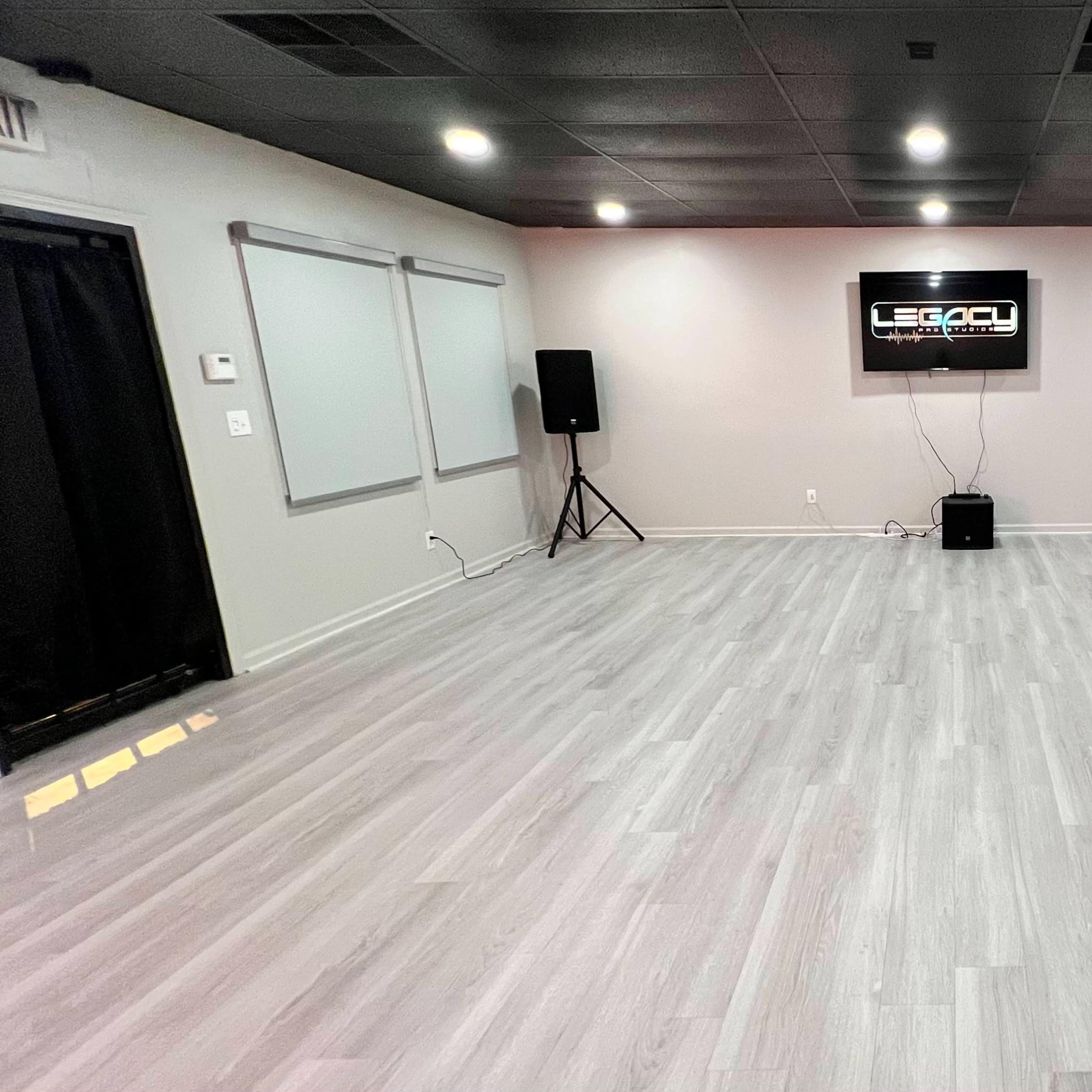 A spacious, empty room with pale wooden flooring, two speakers on stands, and a wall-mounted screen displaying the word "LEGACY."