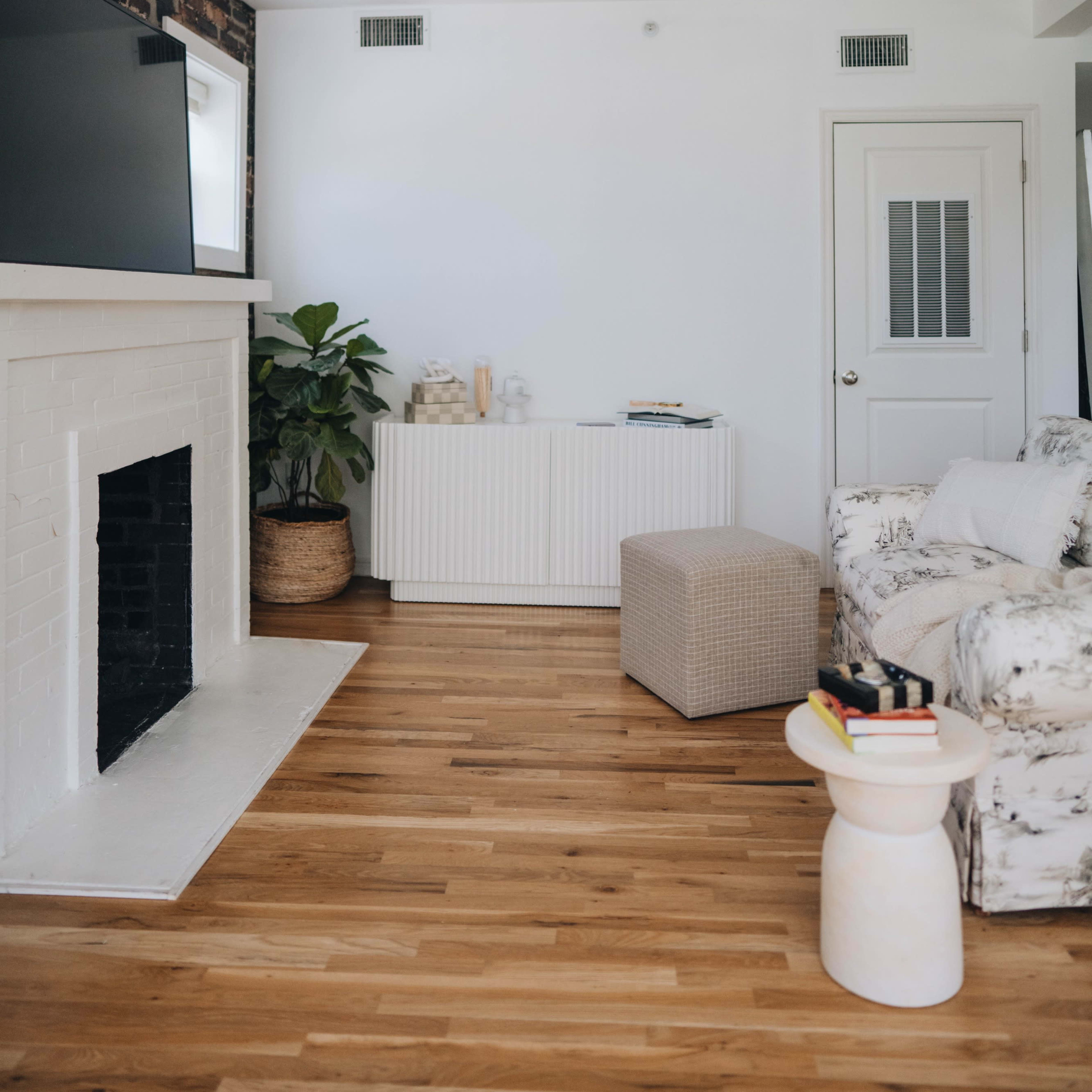 A bright living room features a white brick fireplace, a patterned sofa, a small ottoman, and a large television on the wall.