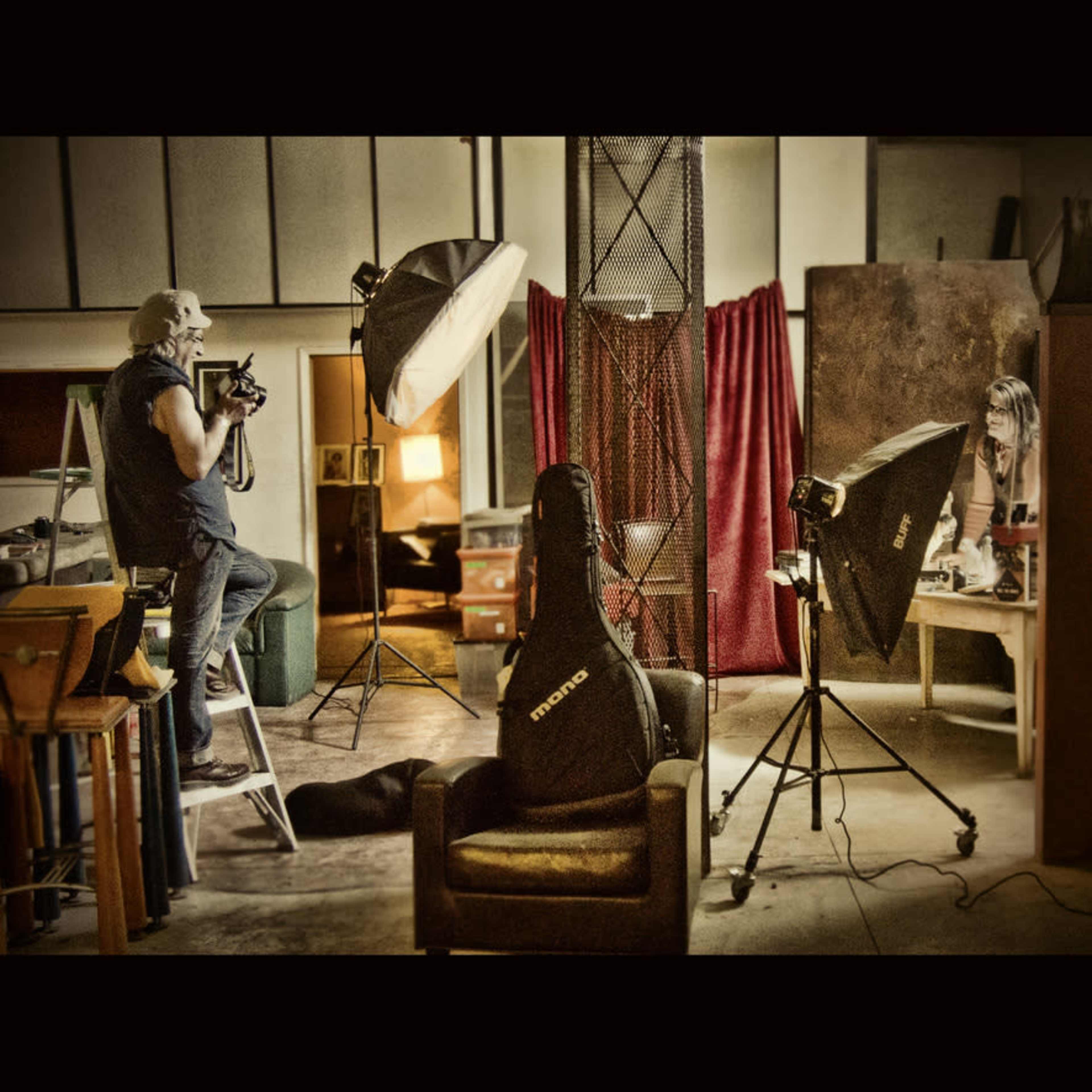 A photographer on a ladder captures an image of a model at a studio, with various lighting equipment and props in the background.