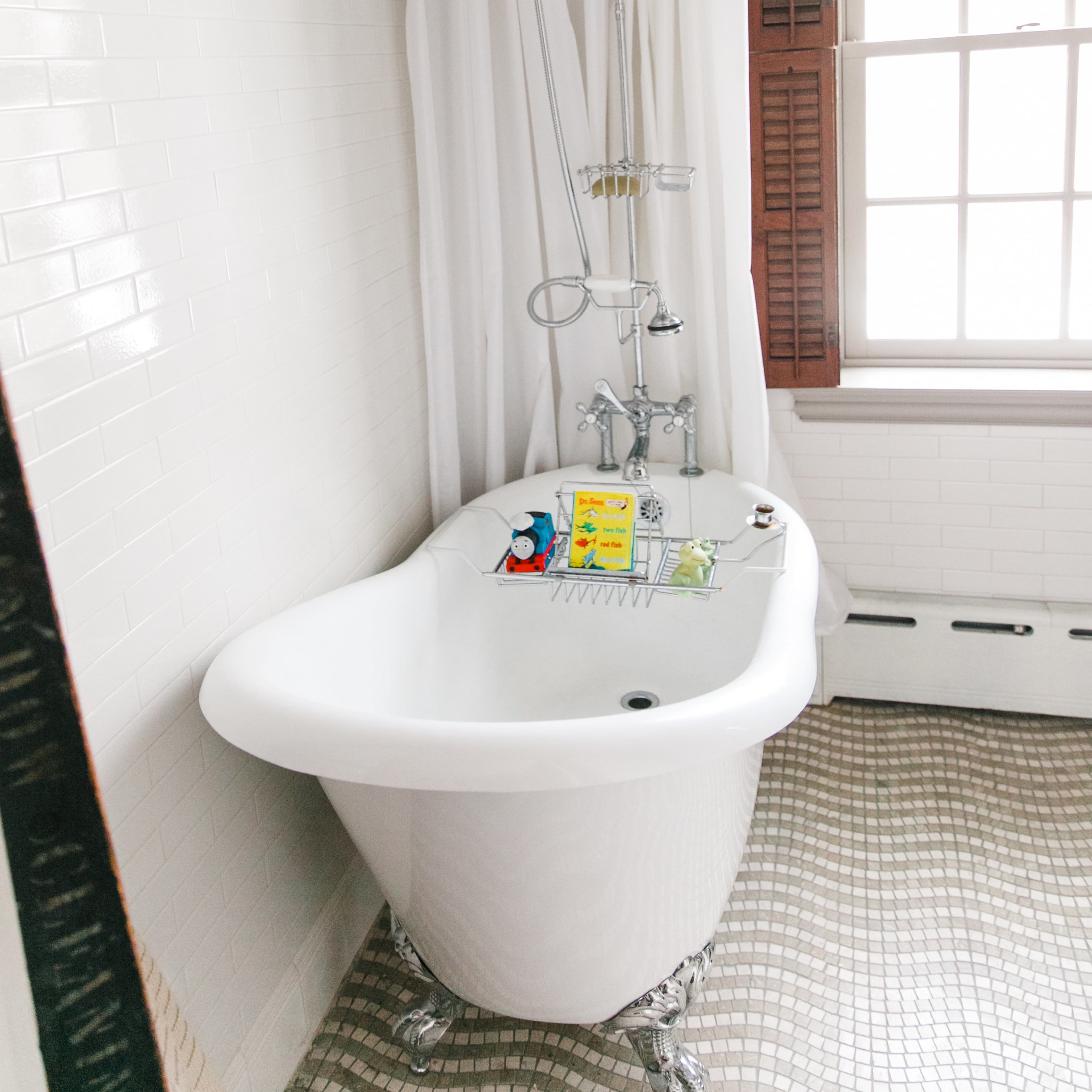 A vintage freestanding bathtub with a shower overhead, surrounded by white tile walls and a window with wooden shutters.