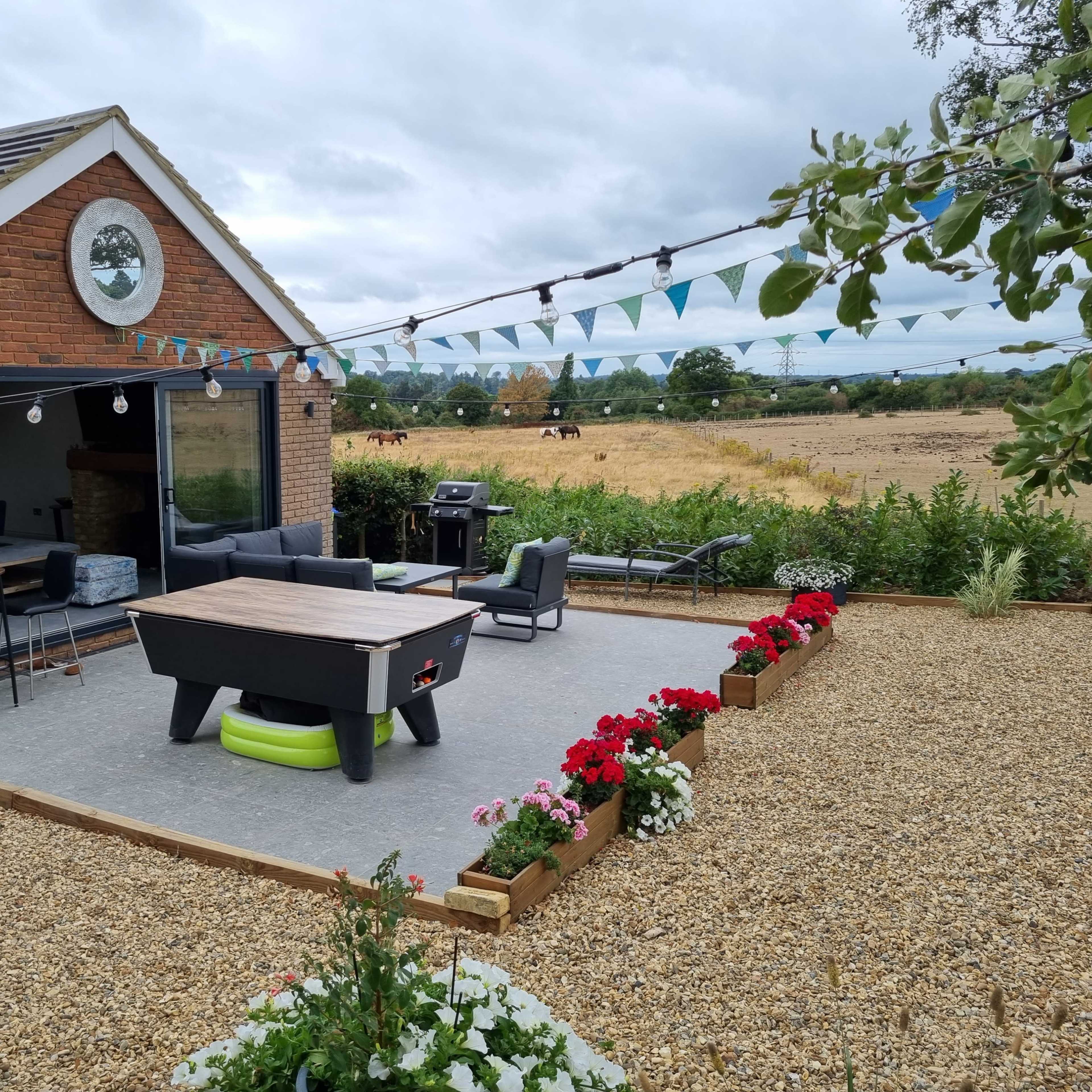 A backyard patio with a pool table and seating area is adjacent to a brick house overlooking a field with grazing horses.