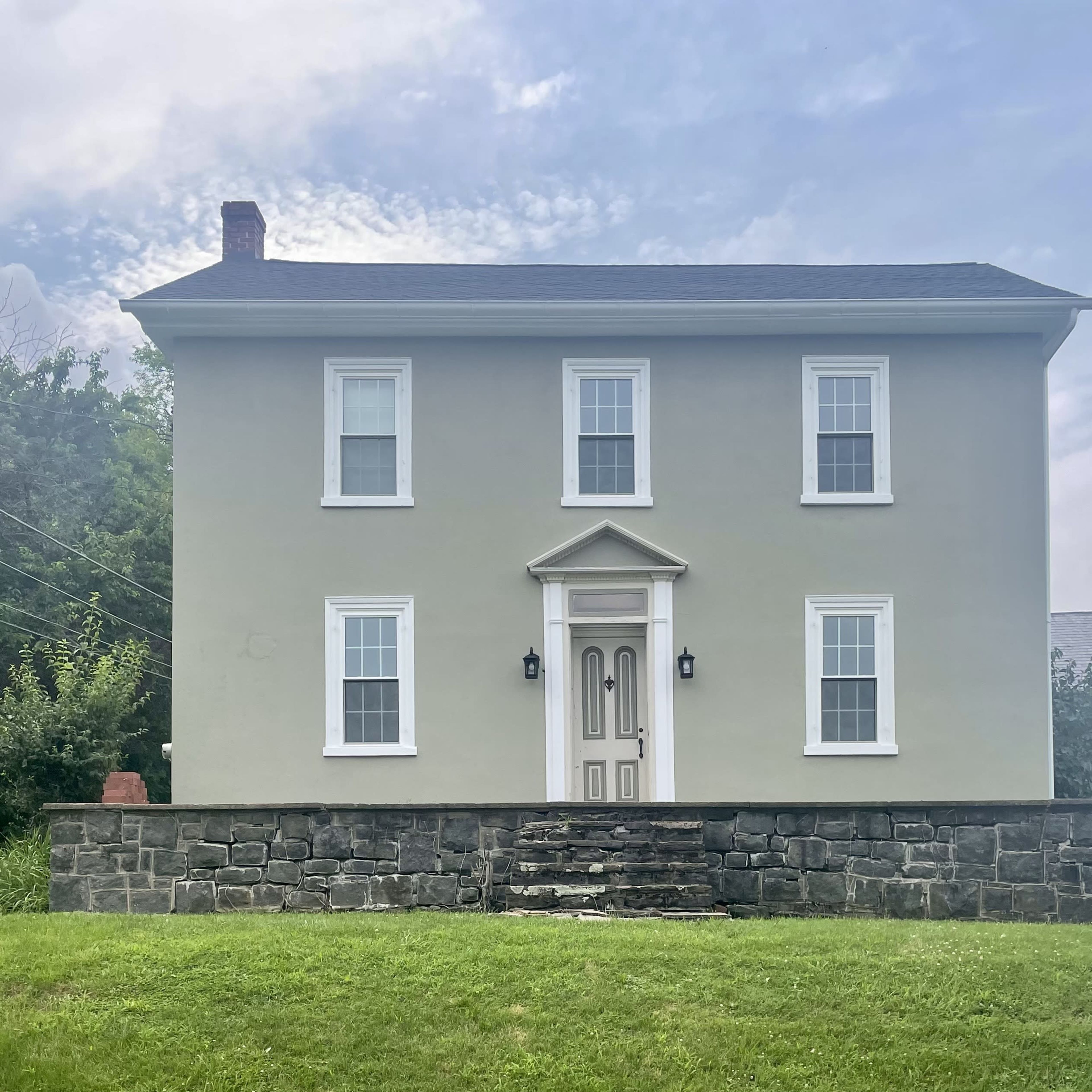 A two-story gray house with white trim and a central entrance stands on a grassy lot, flanked by trees.