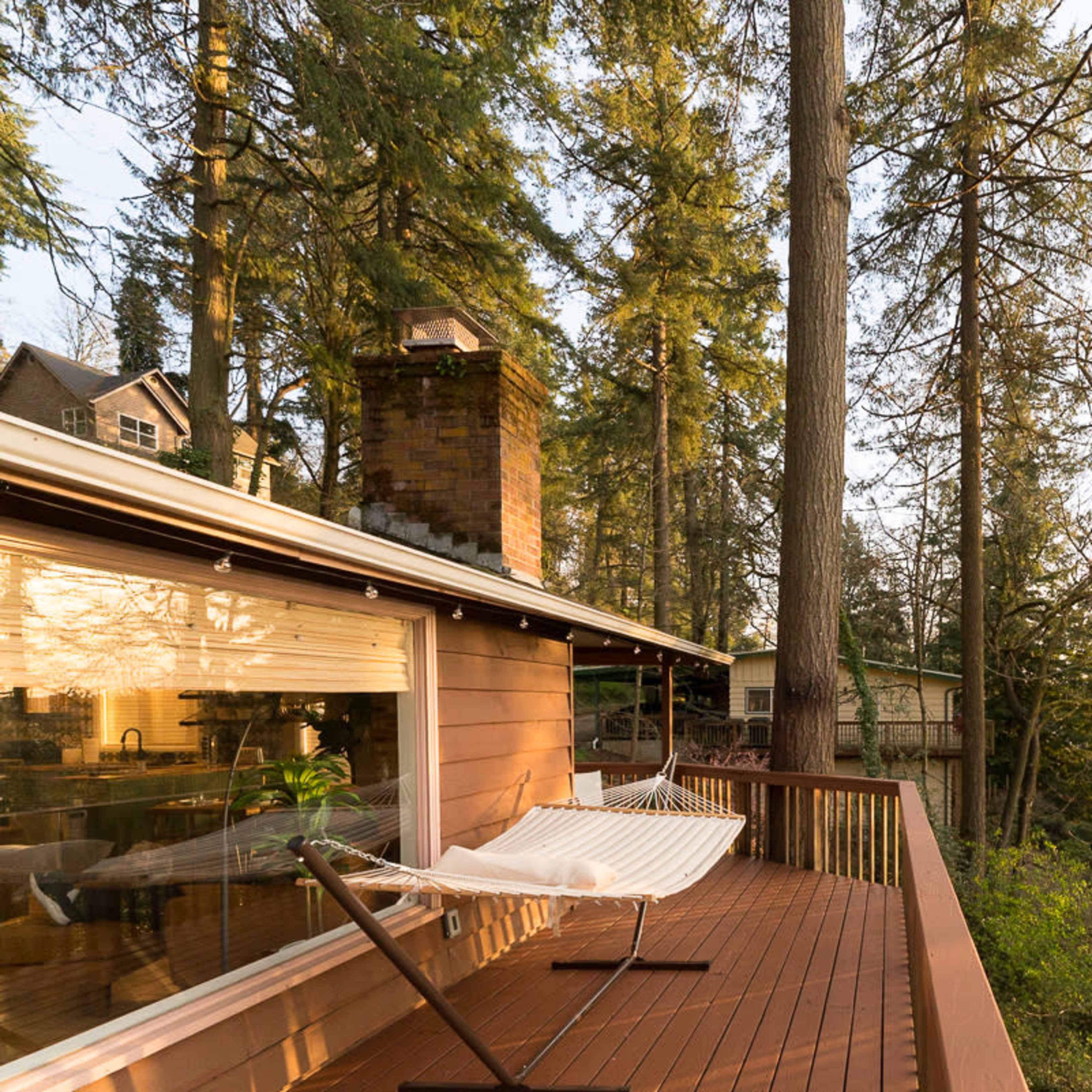 A wooden deck with a hammock overlooks a wooded area and a house partially visible through the trees.