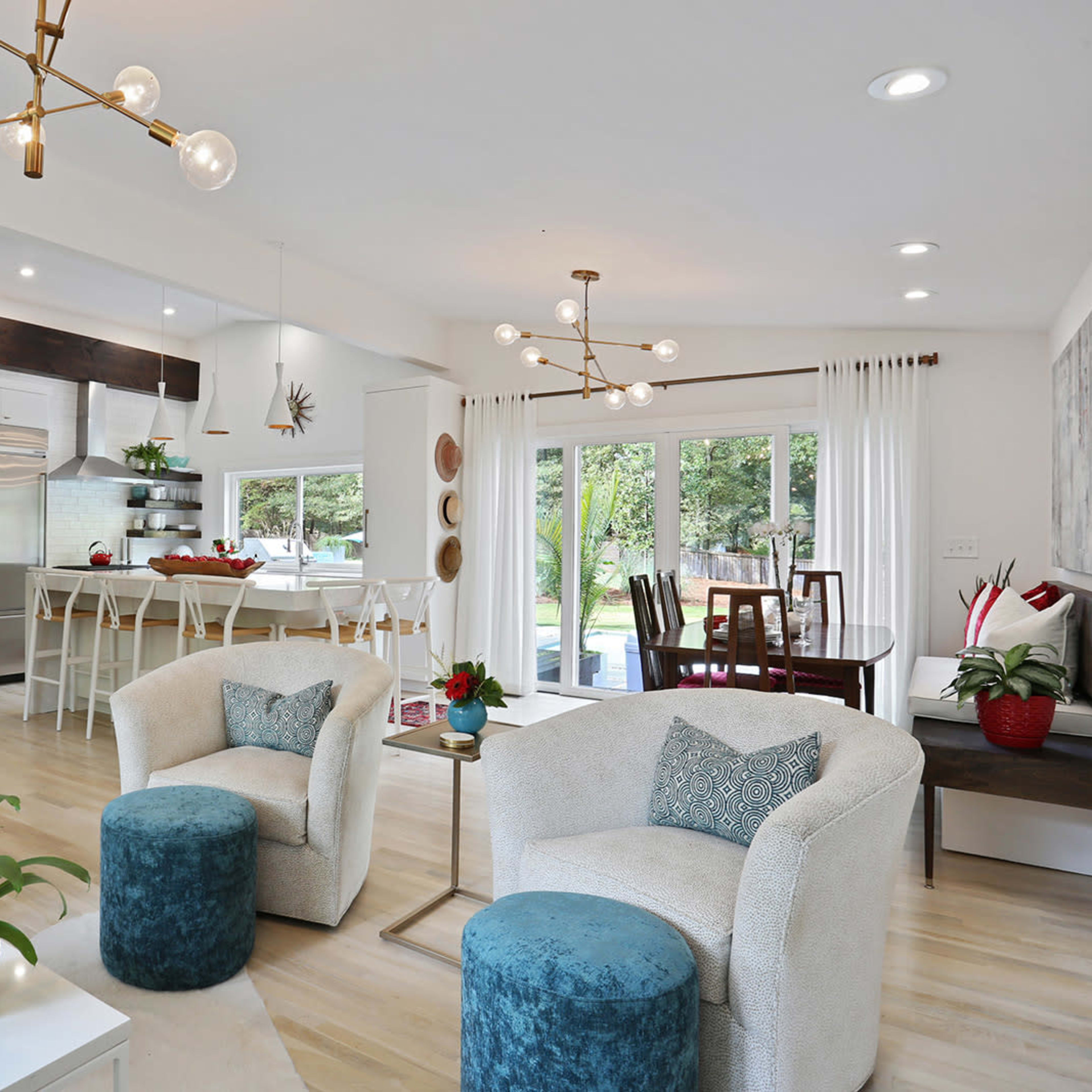 A modern living space features a seating area with light-colored furniture, a dining table in the background, and large windows that open to a view of greenery.