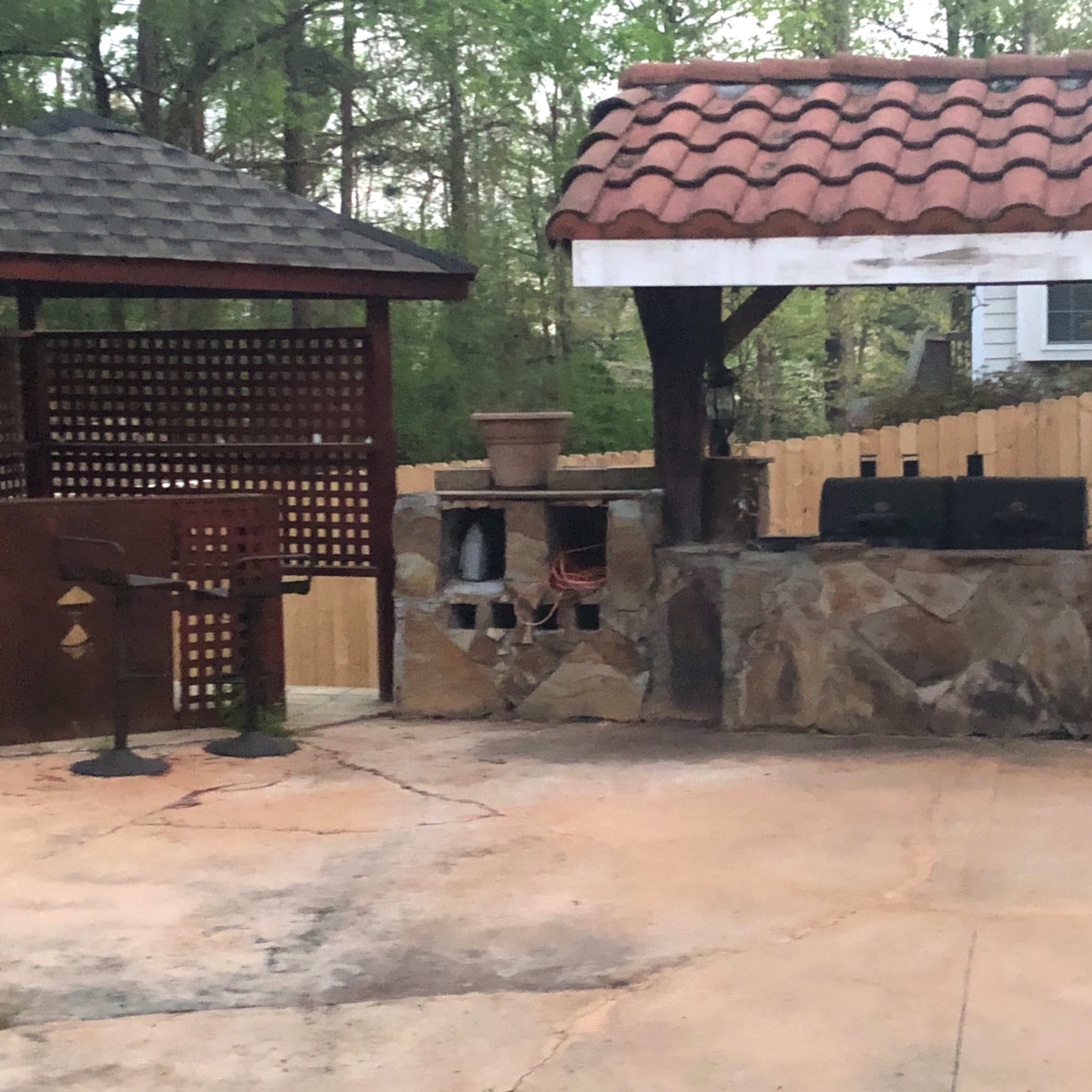 An outdoor patio area with a stone barbecue setup, a wooden gazebo, and a fenced background.