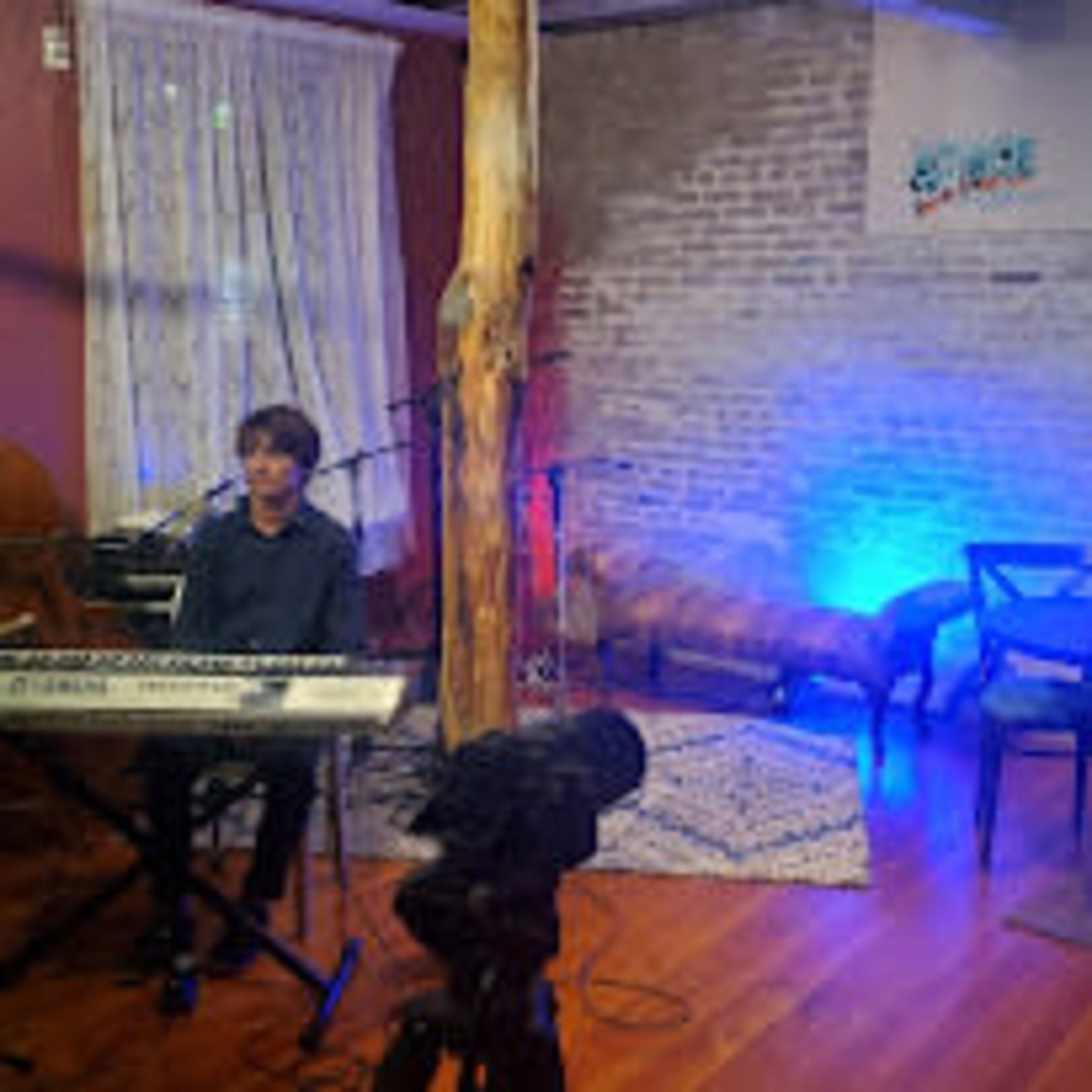 A musician plays a keyboard while a bassist stands nearby, and a woman sits at a table in a room with a rustic ambiance and soft lighting.
