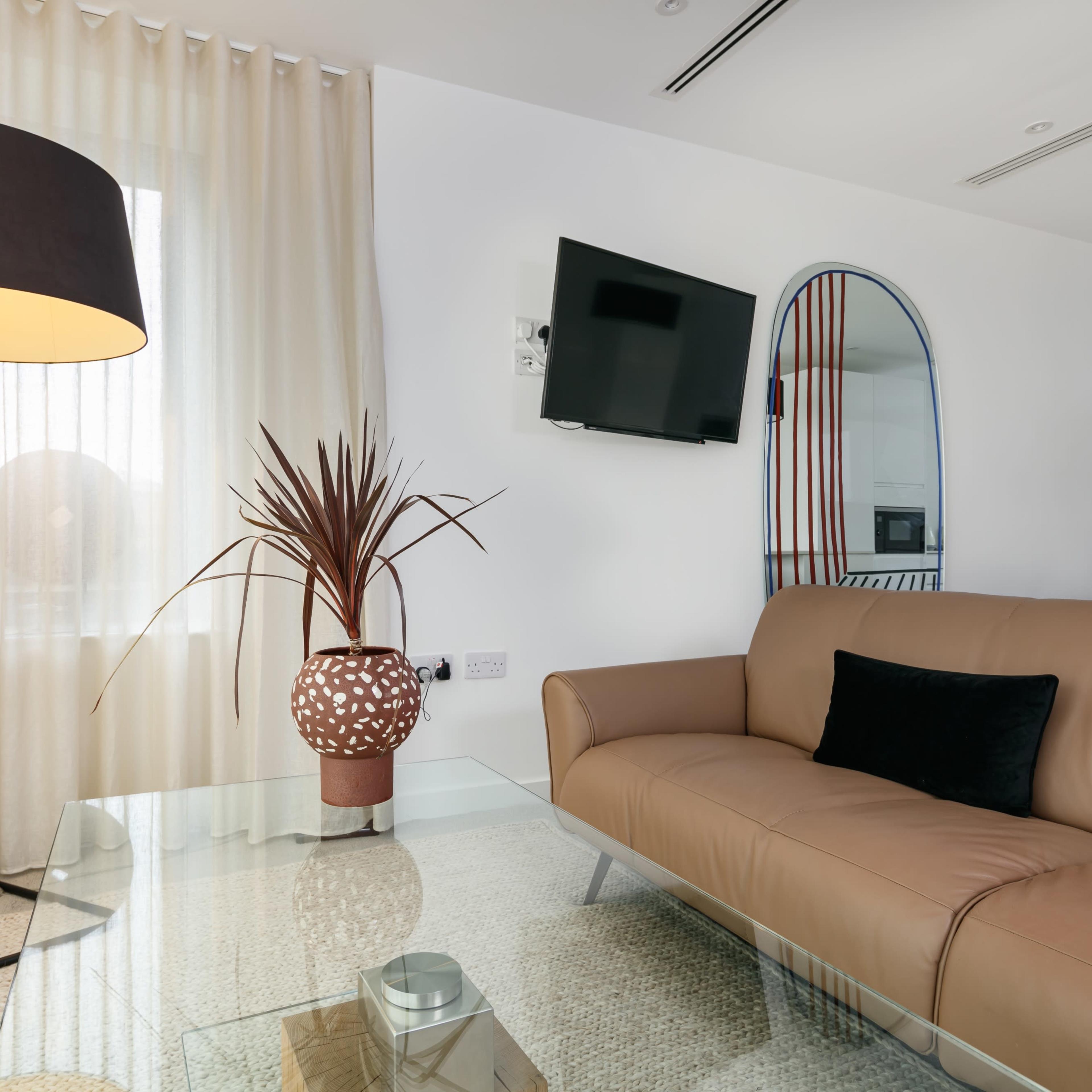 A modern living room features a leather sofa, a glass coffee table, a tall lamp, and a decorative plant beside sheer curtains and a wall-mounted TV.