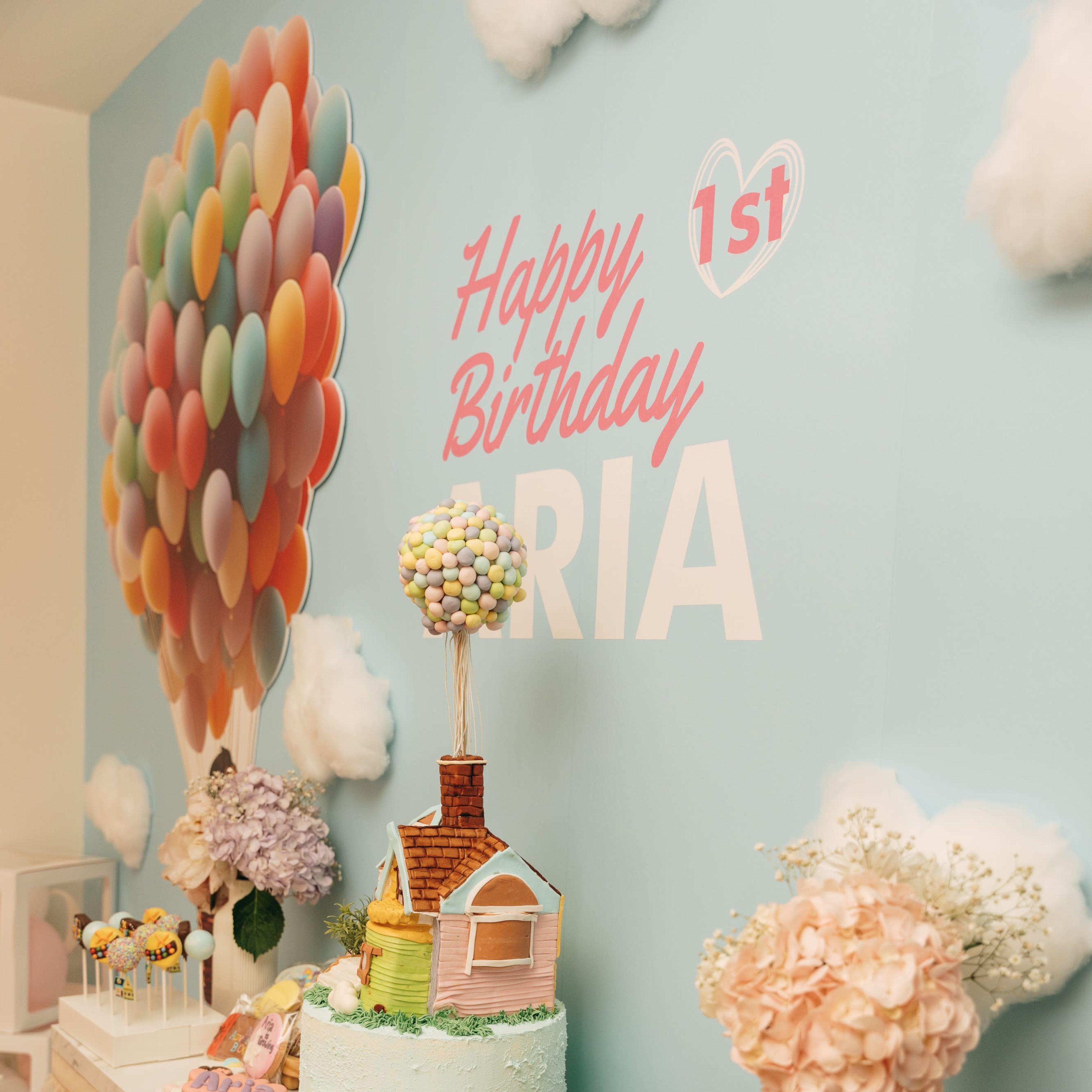 A birthday cake and a variety of desserts are displayed on a table under a colorful balloon-themed backdrop for a first birthday celebration.