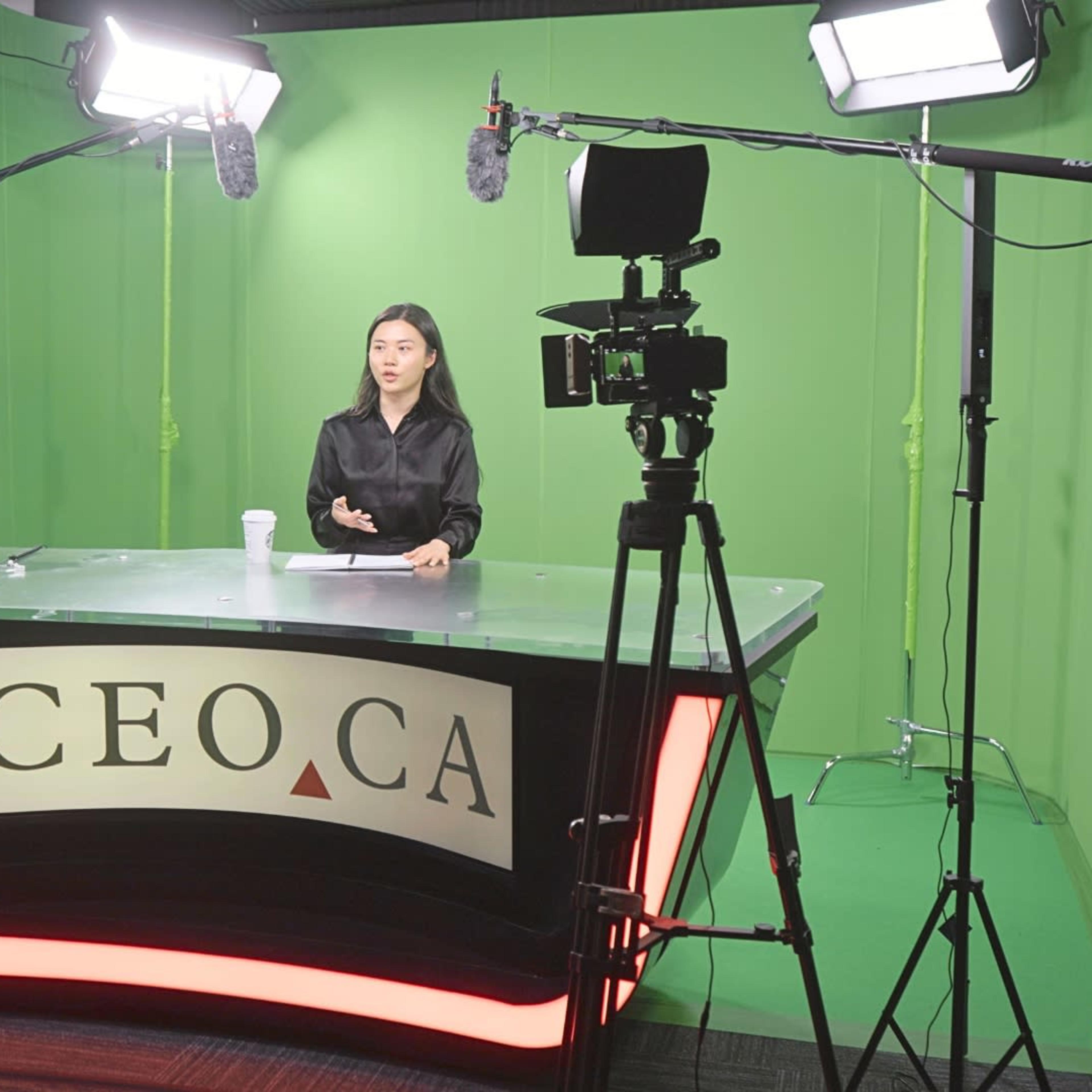 A woman sits at a glass desk with the letters "CEO CA" in front of her, preparing for a presentation in a studio with a green screen backdrop and multiple camera setups.