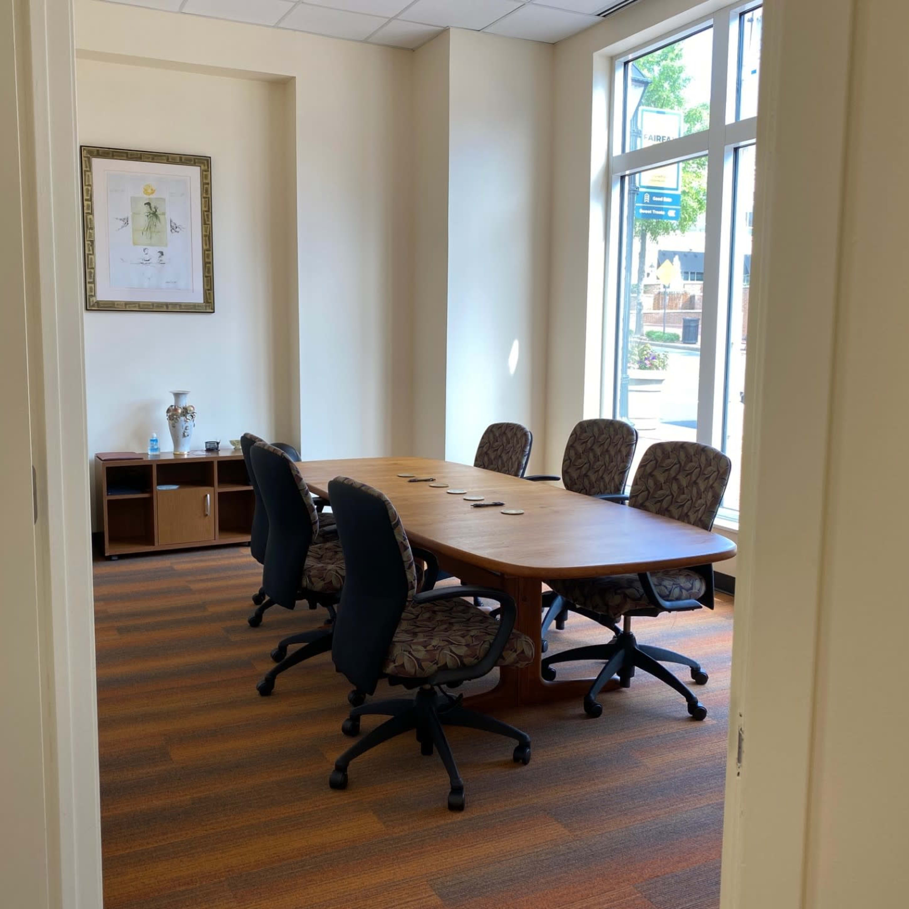 A vacant conference room features a large wooden table surrounded by five office chairs, with a framed artwork on the wall.