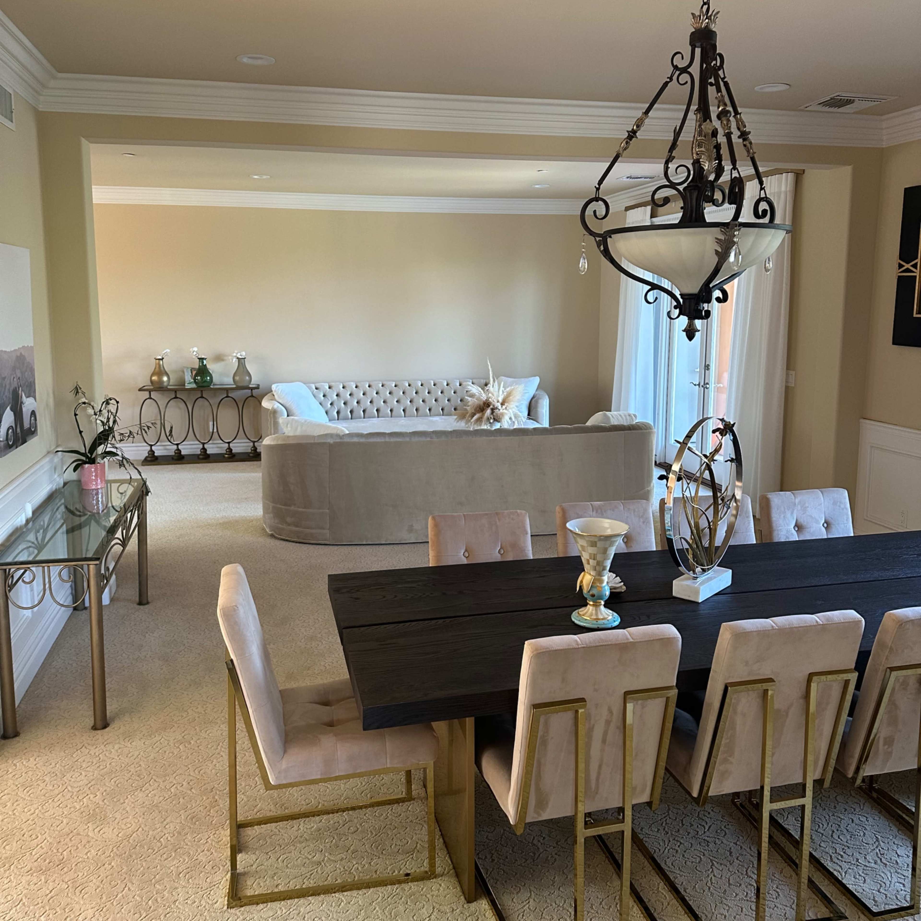 A dining area features a long black table with gold-accented chairs, adjacent to a spacious living room with a light-colored sofa.