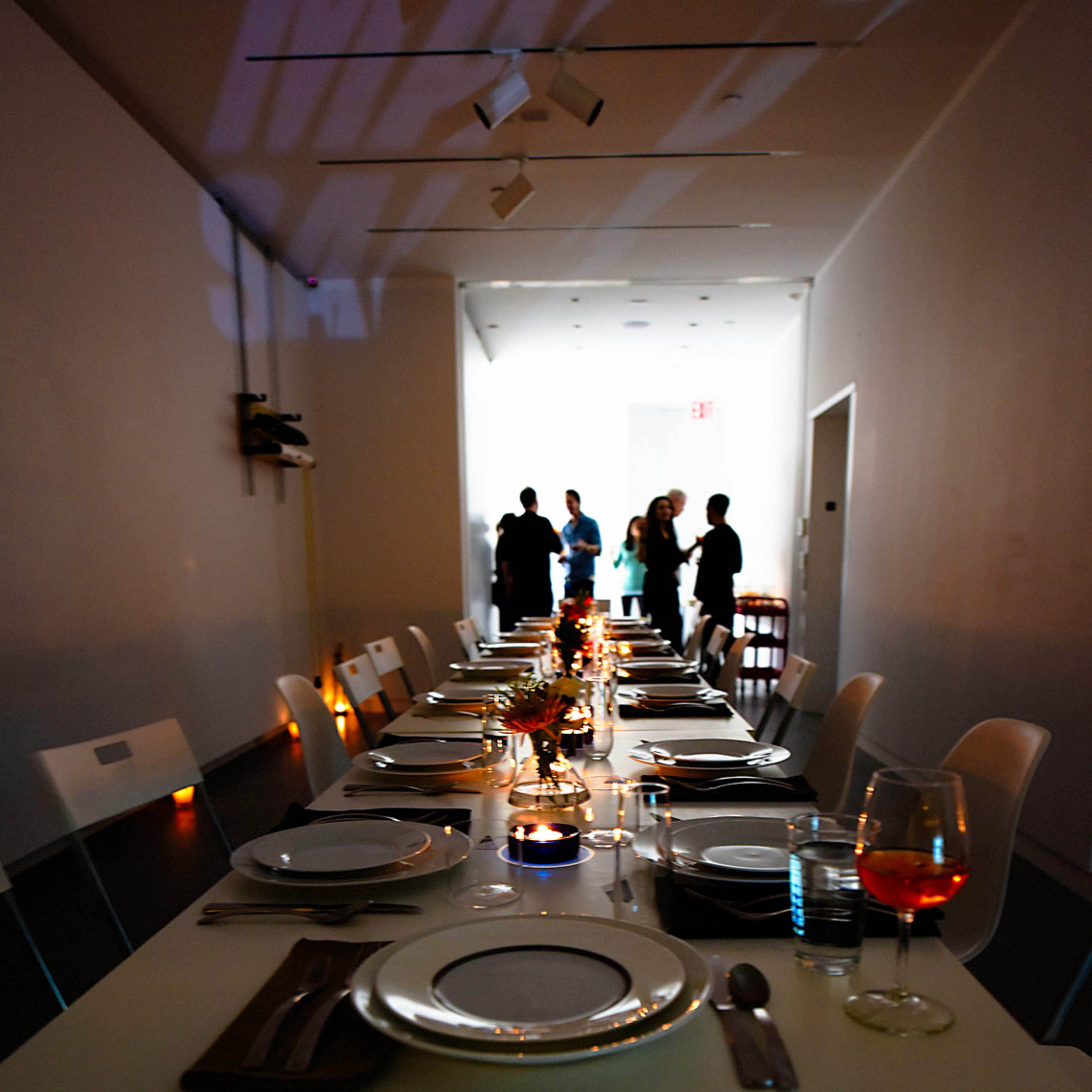 A long dining table is elegantly set with plates and glassware, while people gather in the background of a well-lit room.