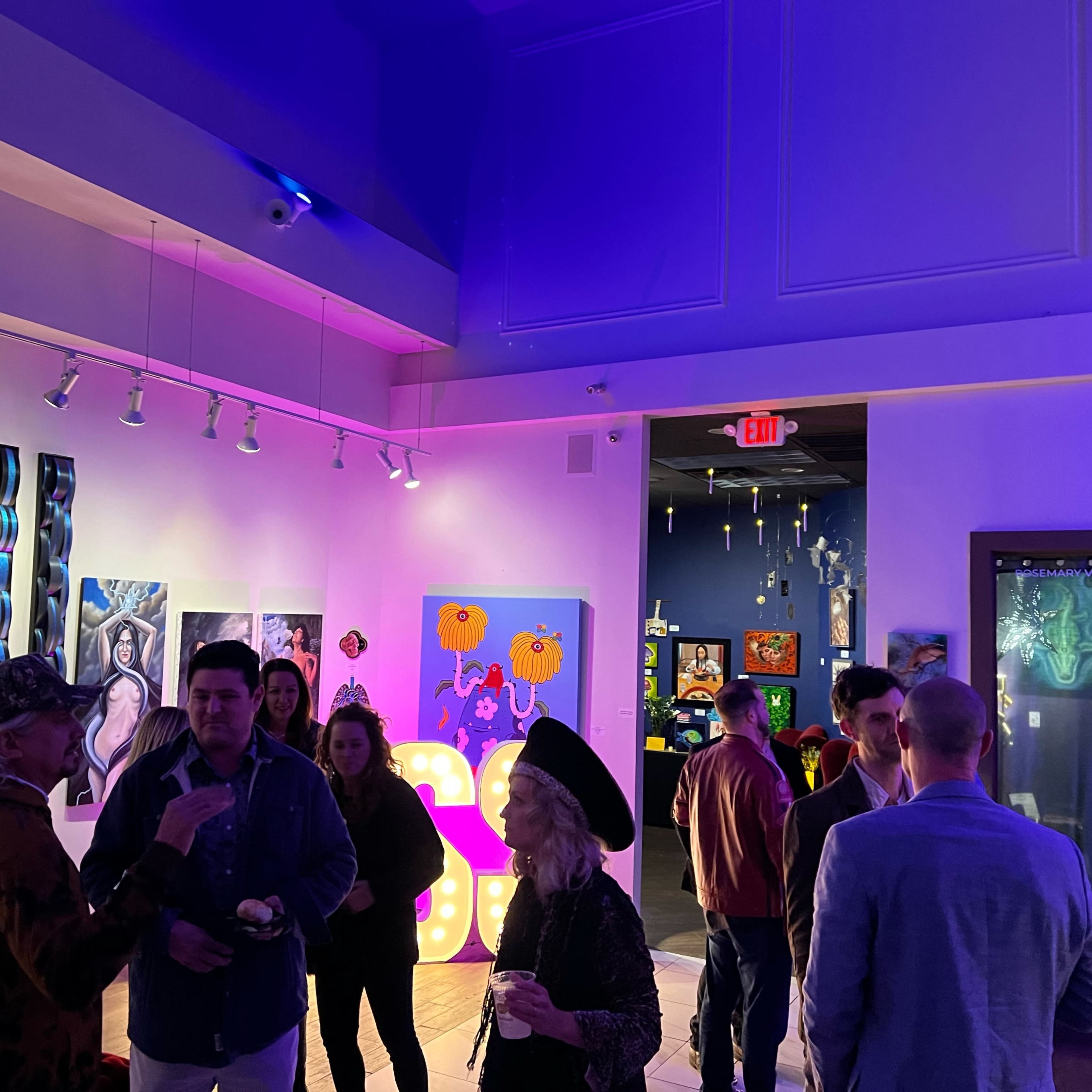 People socialize during an art exhibition featuring colorful artwork and vibrant lighting in a gallery space.