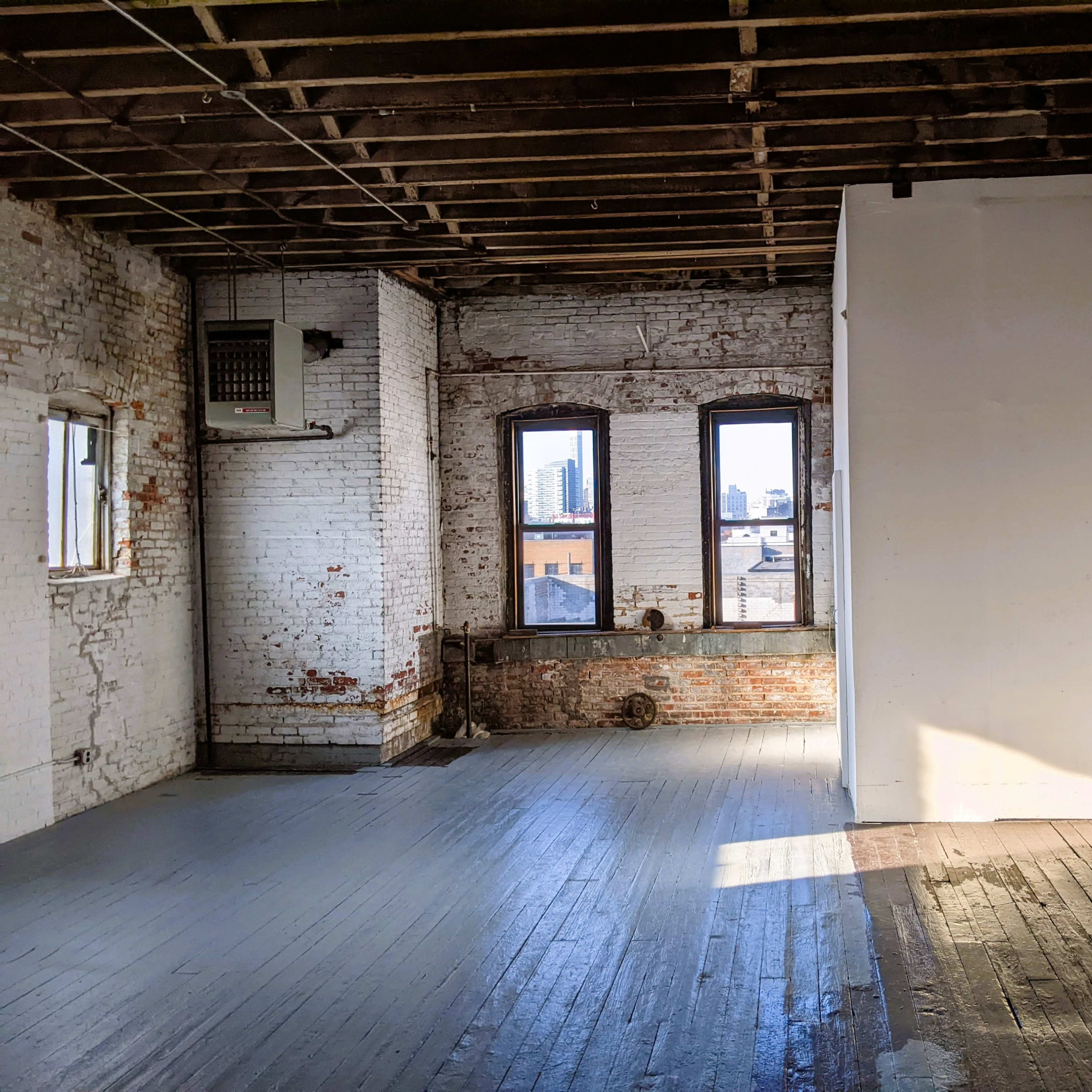 An empty industrial space features exposed brick walls, large windows with a city view, and wooden flooring.