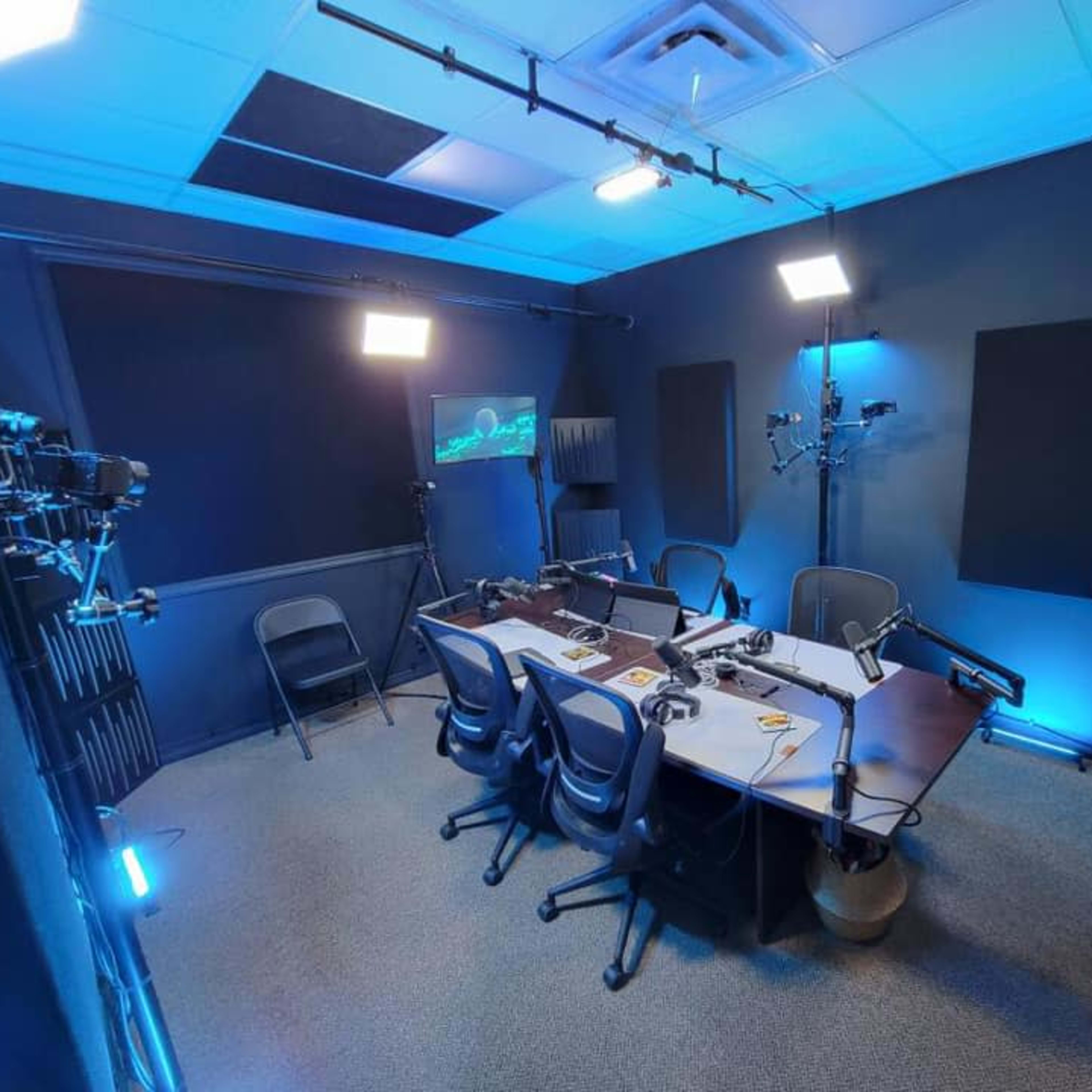 A well-equipped podcast studio with a central table, multiple microphones, and lighting setups in a dark blue-themed room.