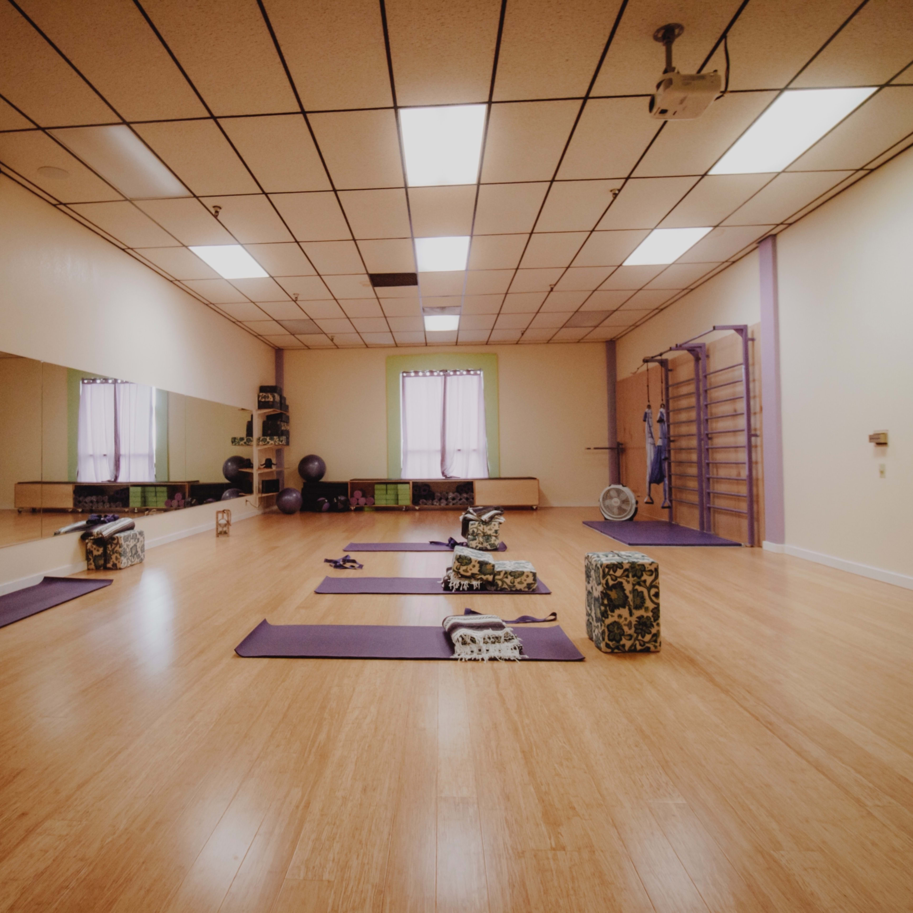 A spacious yoga studio with wooden flooring, yoga mats arranged on the floor, and equipment such as blocks and straps positioned around the room.