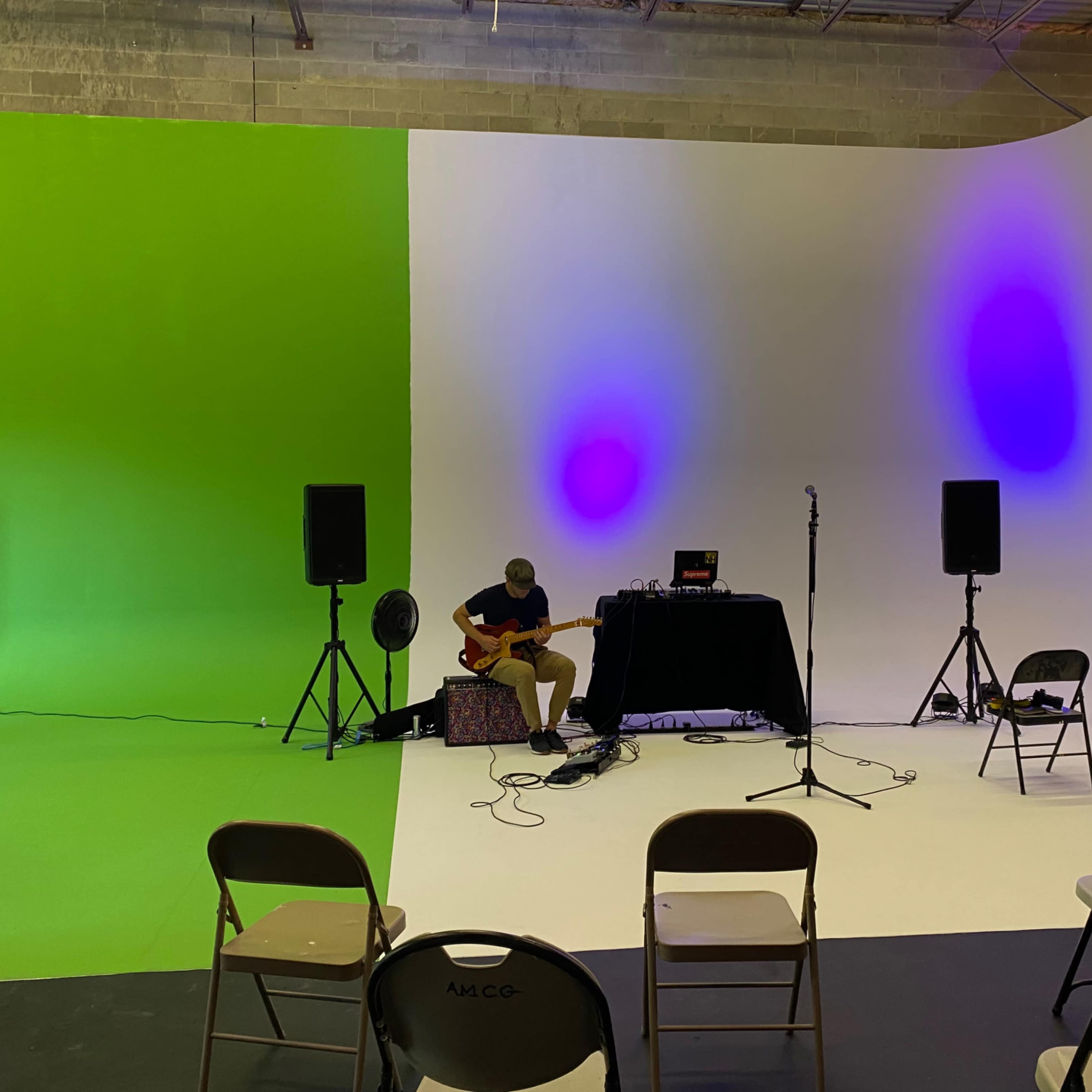 A musician plays guitar on stage with a sound setup in front of a backdrop that is divided into green and white sections, with speakers and chairs arranged for an audience.