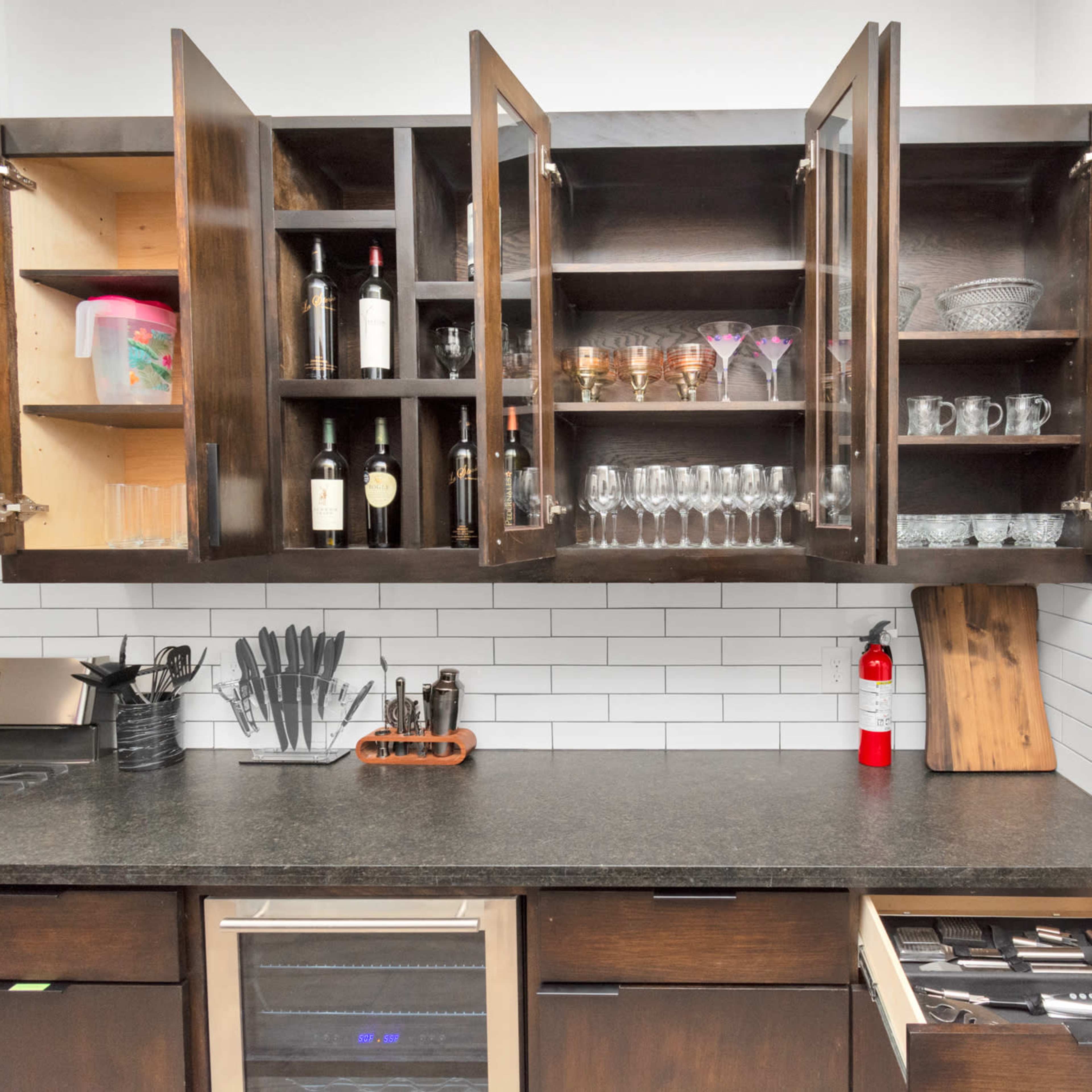 A kitchen with open cabinets displaying glassware and bottles, a countertop with a knife block, and a partially opened drawer revealing utensils.