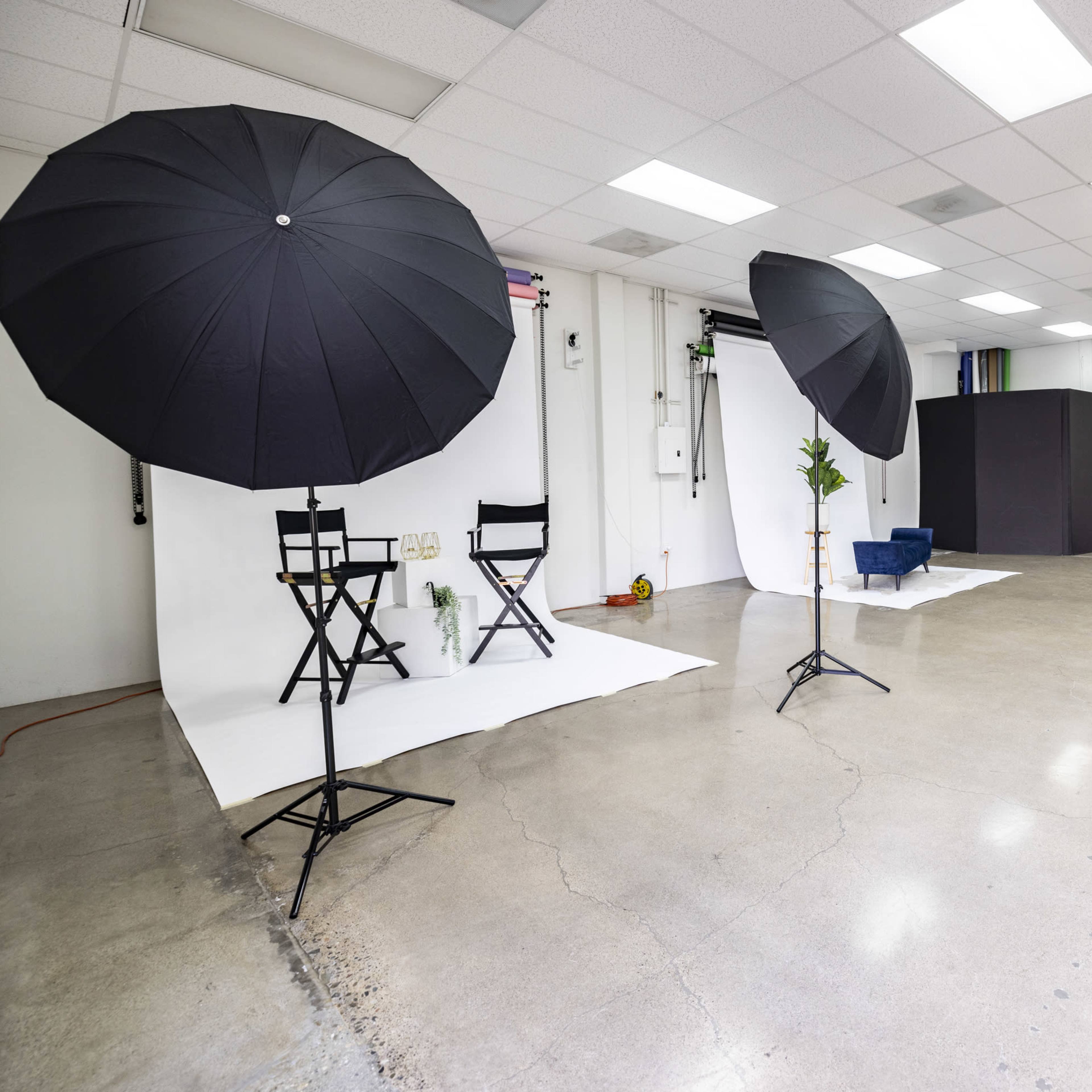 A spacious studio with two black umbrellas set up beside two director's chairs on a white backdrop, and a dark box in the background against a minimalistic interior.