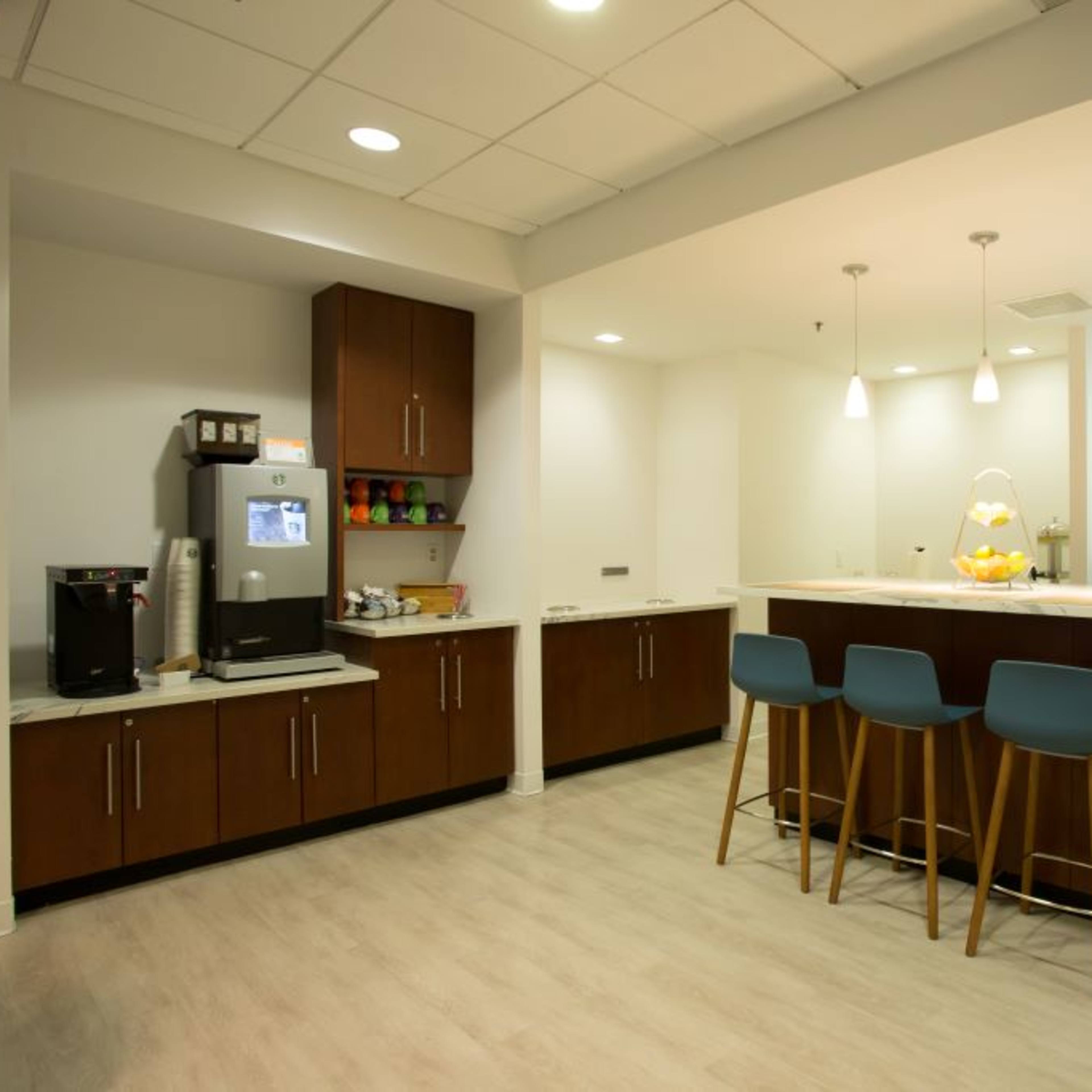 A modern kitchen features a central island with three stools, cabinetry, a refrigerator, and various appliances along the walls.