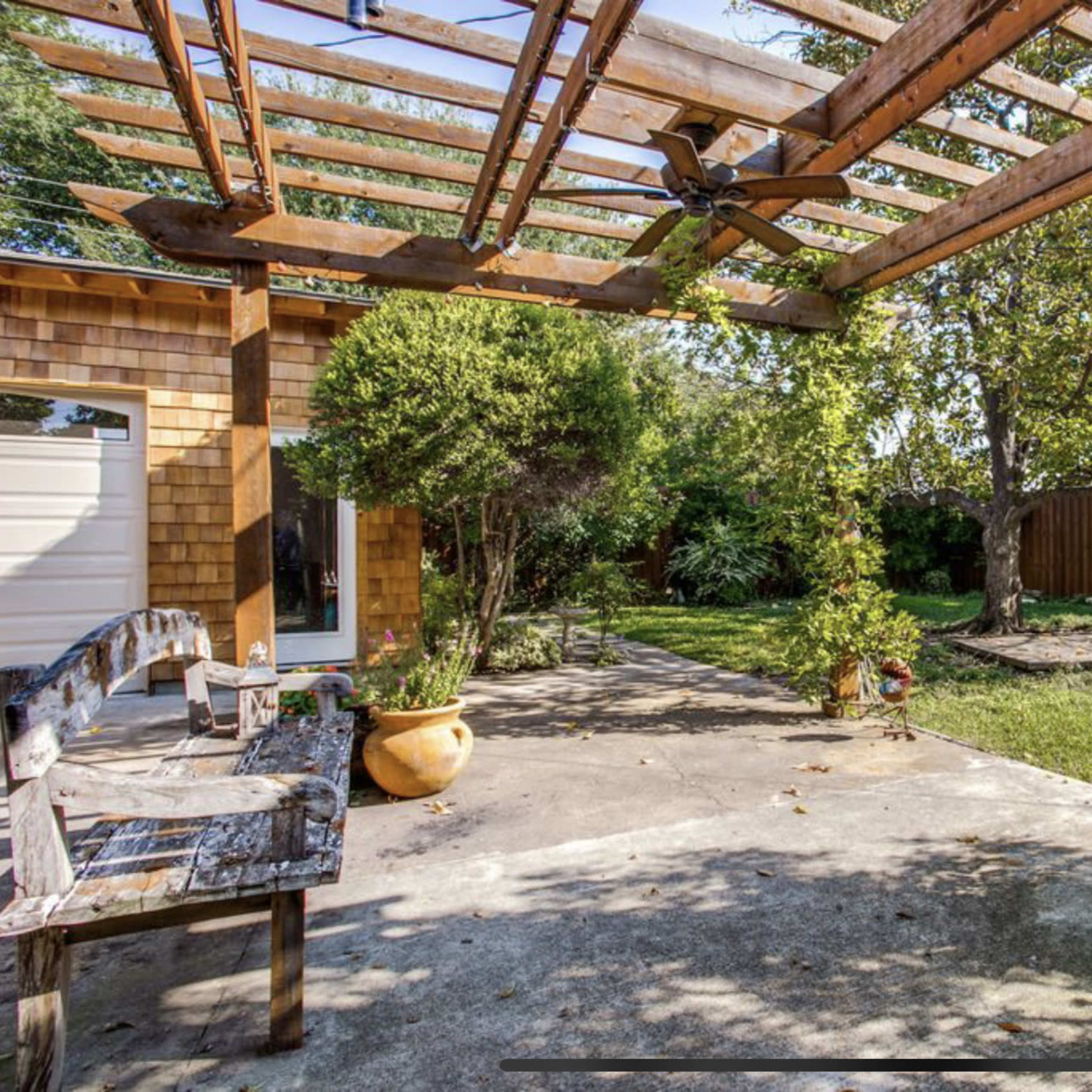 An outdoor space features a wooden pergola with climbing plants, a stone pathway, and greenery in the background.