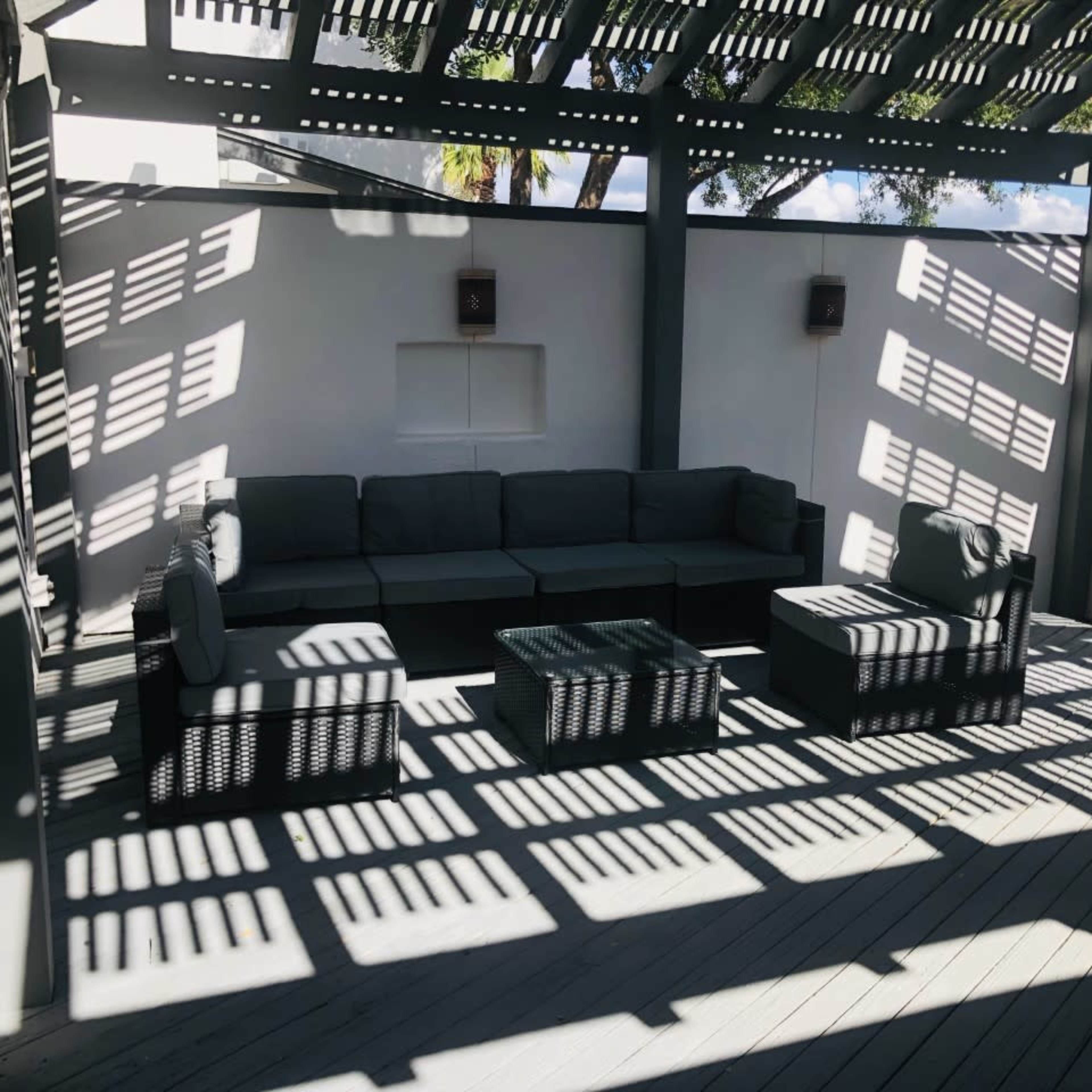A shaded outdoor seating area features a sectional couch and coffee tables arranged on a wooden deck.