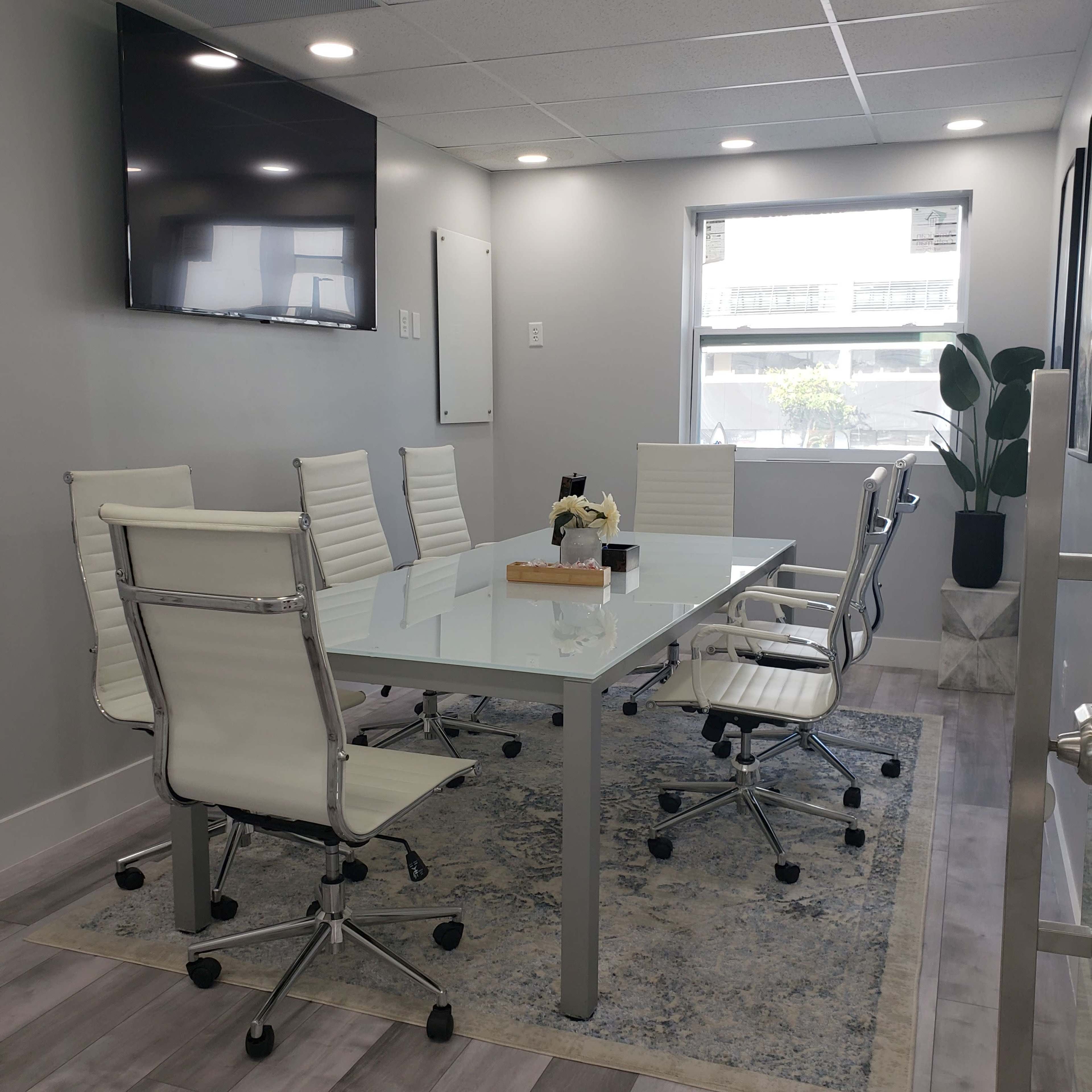 A sleek conference room features a glass table surrounded by white chairs, a large wall-mounted TV, and a potted plant.