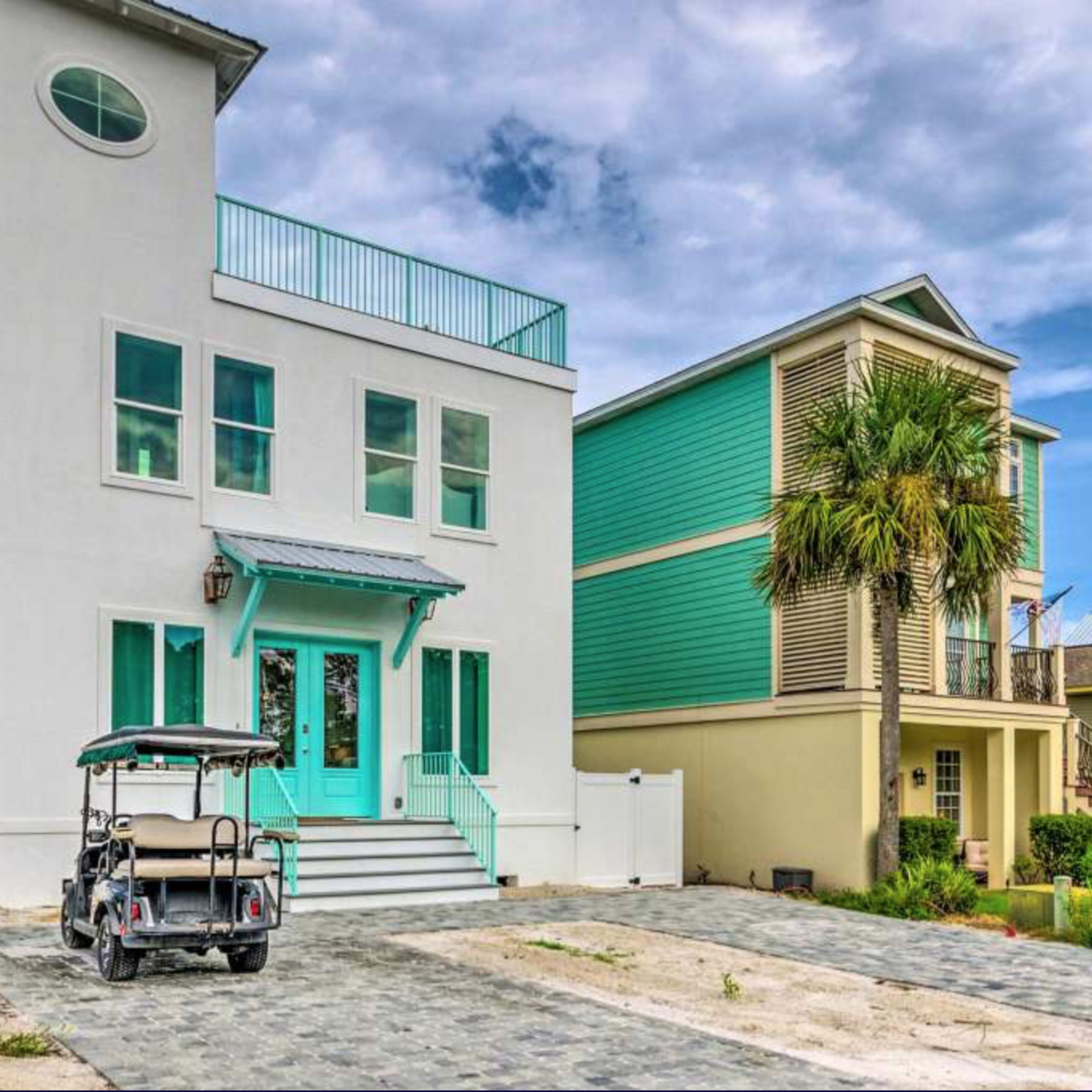 A modern three-story house with teal accents and a golf cart parked in front is surrounded by a landscaped yard and a wooden fence.
